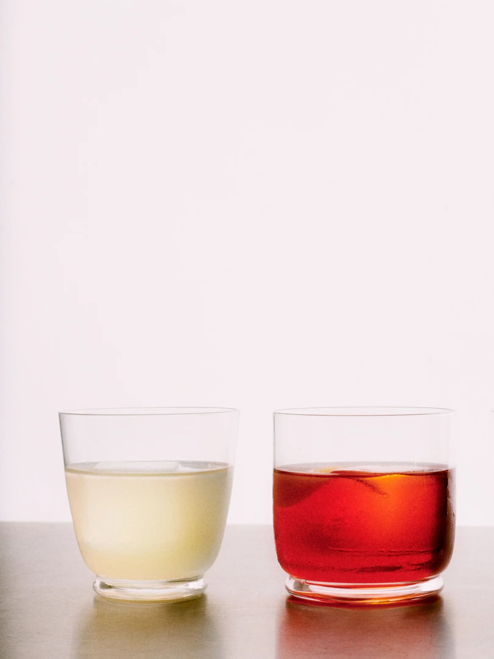 Two clear tumblers from the Walter 01 - Tumbler Set by Service Projects are filled with different beverages on a plain surface against a white background. The left tumbler contains a light yellow liquid, and the right tumbler holds a red liquid, both appearing to be non-carbonated drinks. Each piece showcases exquisite mouth-blown glassware artistry.