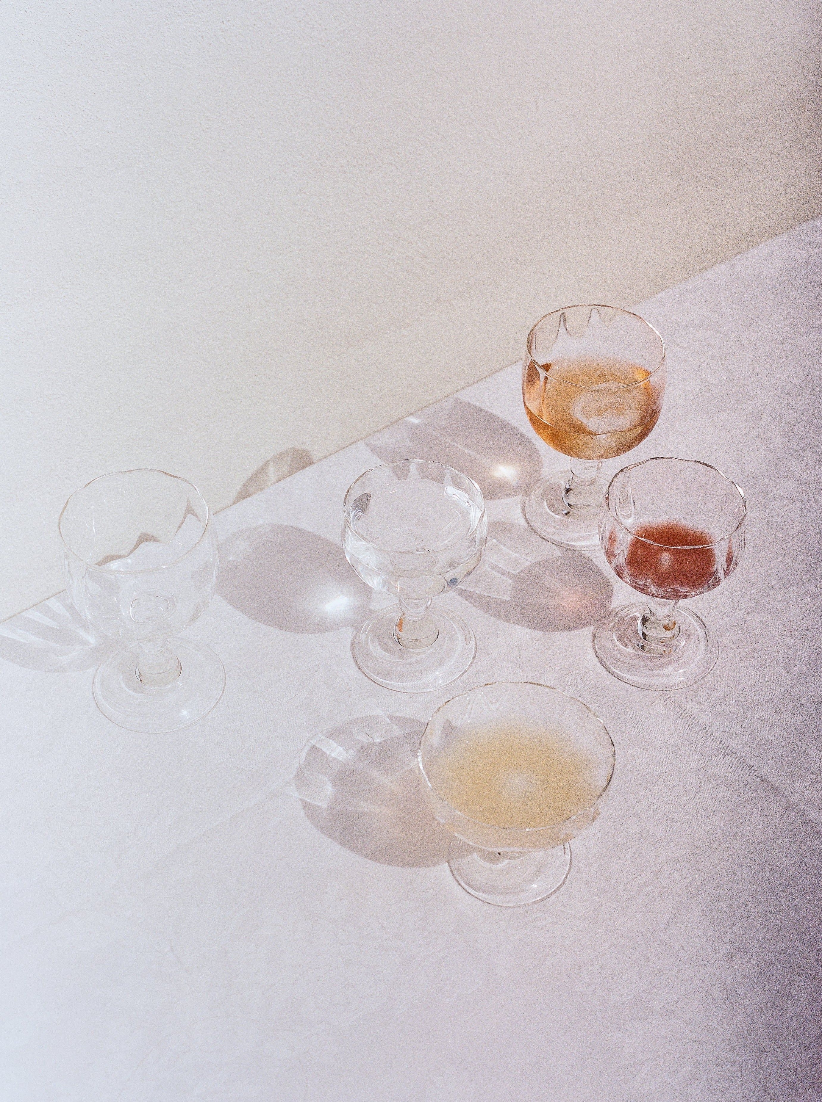 Four elegant Akua Objects Alban Wine Glasses with varying amounts of liquid, arranged on a textured white tablecloth under soft lighting, casting gentle shadows.