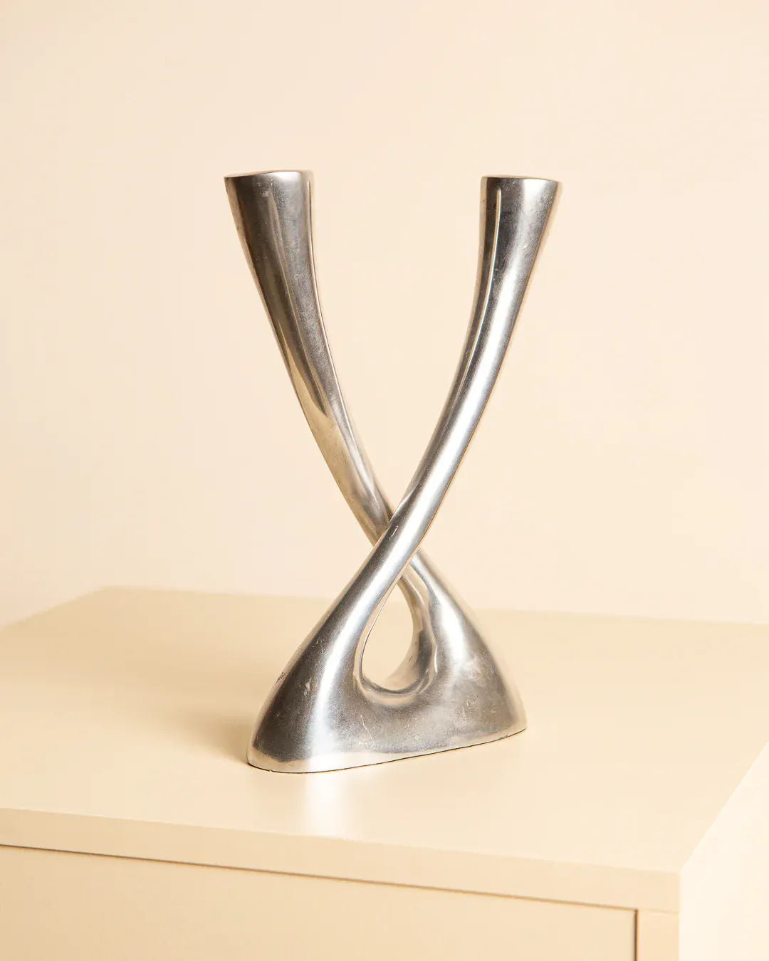 The Aluminum "Croisé" Candleholder 80's by Treaptyque is a modern centerpiece with timeless allure, featuring two intertwined and elongated forms crafted from silver metal. It stands gracefully on a light-colored surface, boasting smooth, reflective curves in an abstract design set against a neutral backdrop.