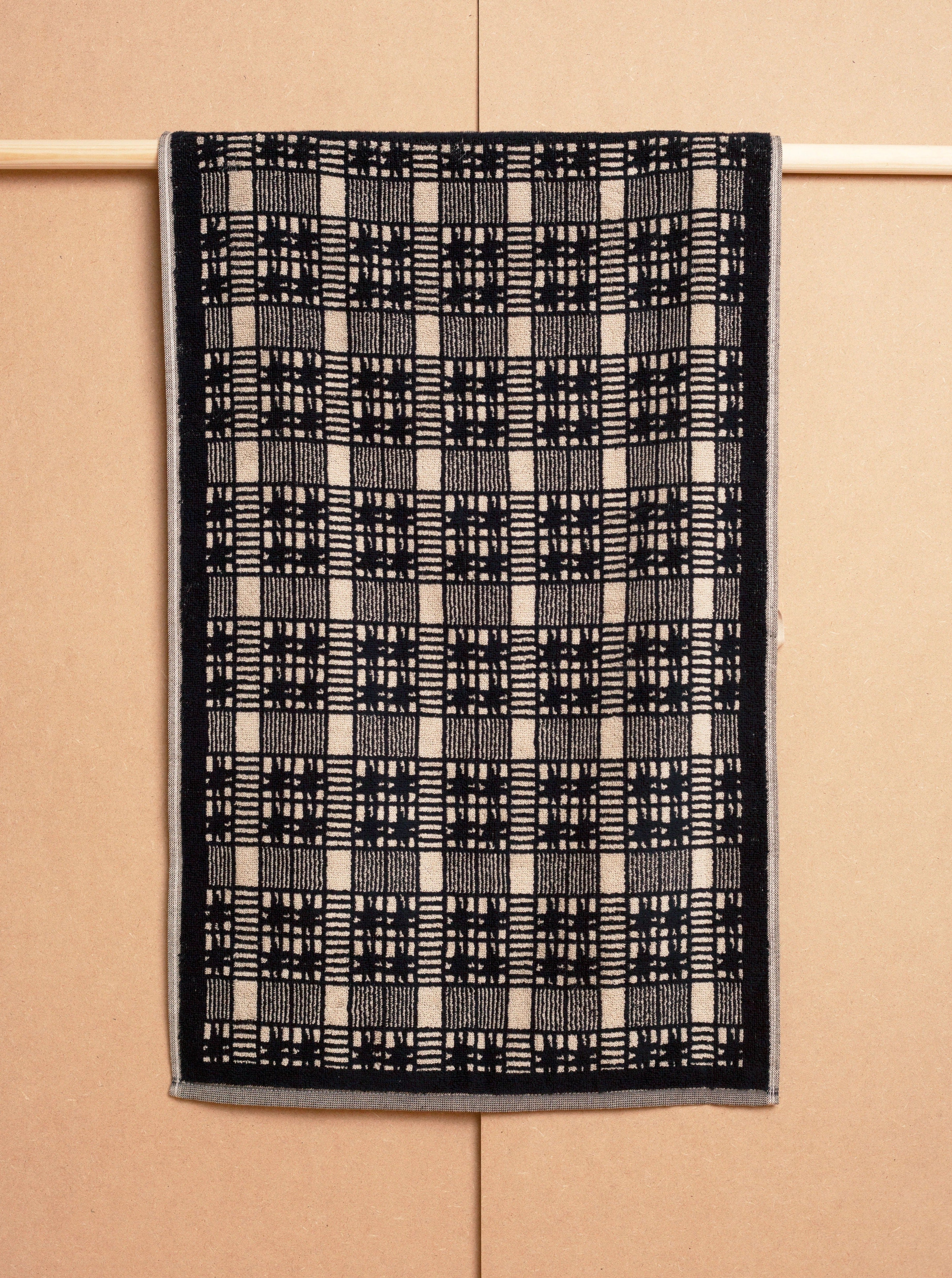 A black and white two-tone checkered design Alma Hand Towel hanging against a beige cardboard background, featuring a geometric pattern with varying grid-like patterns by Autumn Sonata.