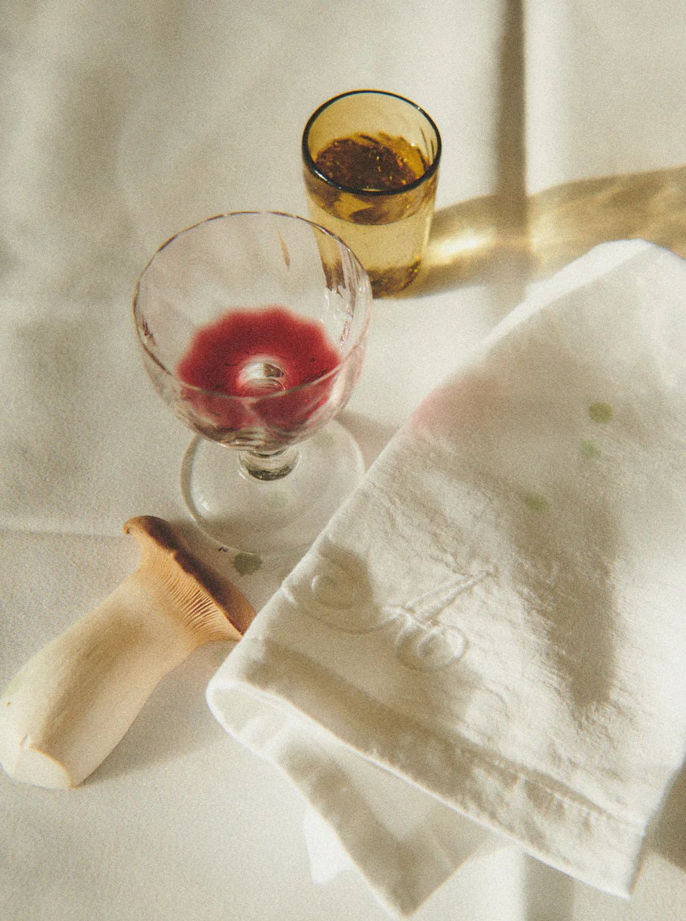 A textured image featuring a table setting with a glass of red wine, a yellow Akua Objects Alban Wine Glass - Set of Four half-filled, and a stained white napkin beside brass cutlery, all bathed in