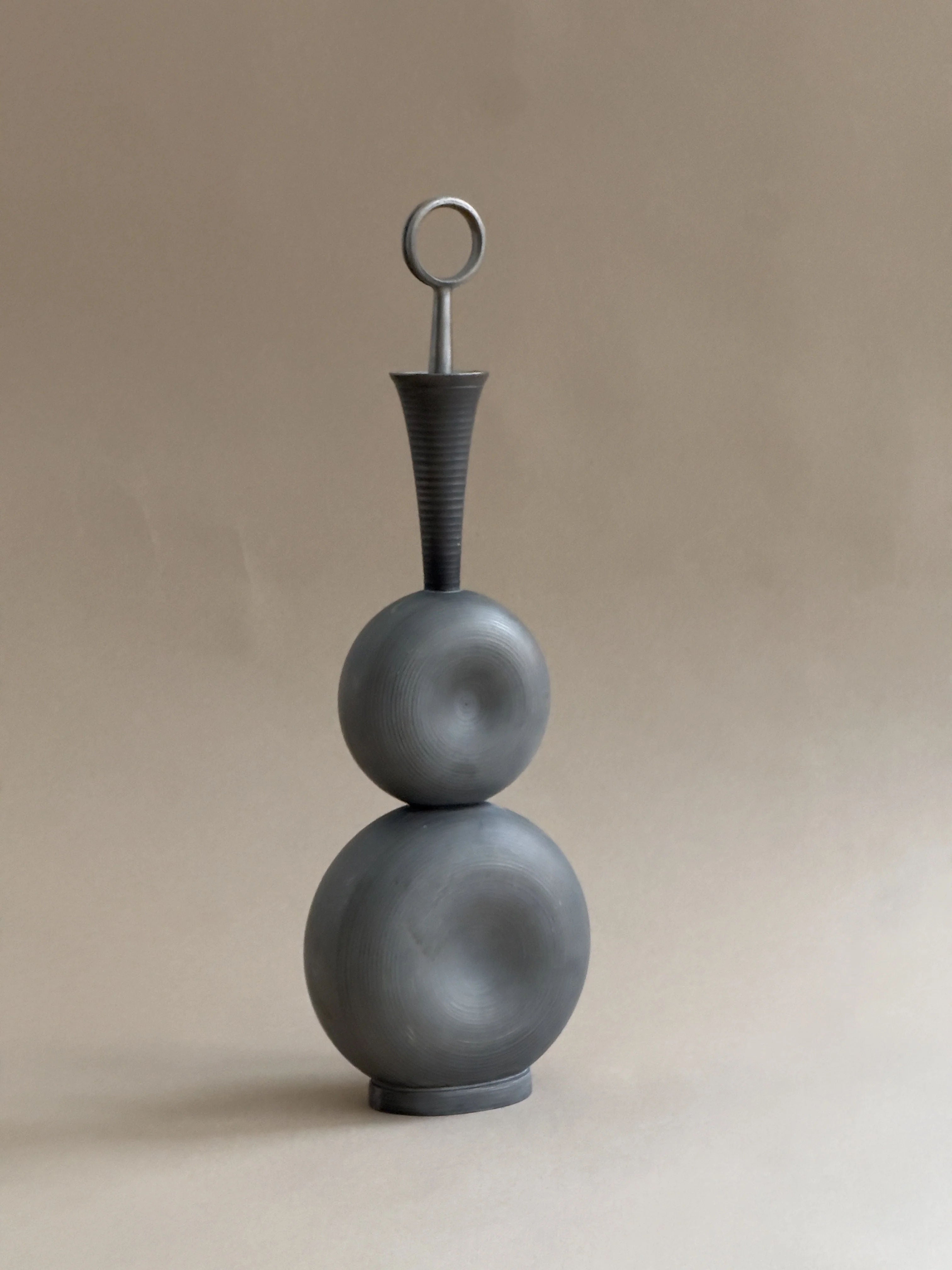 The Modernist Pewter Decanter by Galerie Storm is a matte gray sculpture with a round base and orb, leading to a conical neck with a circular handle. Crafted in pewter and echoing Gunnar Havstad's style, it exudes minimalist elegance against a soft beige backdrop.