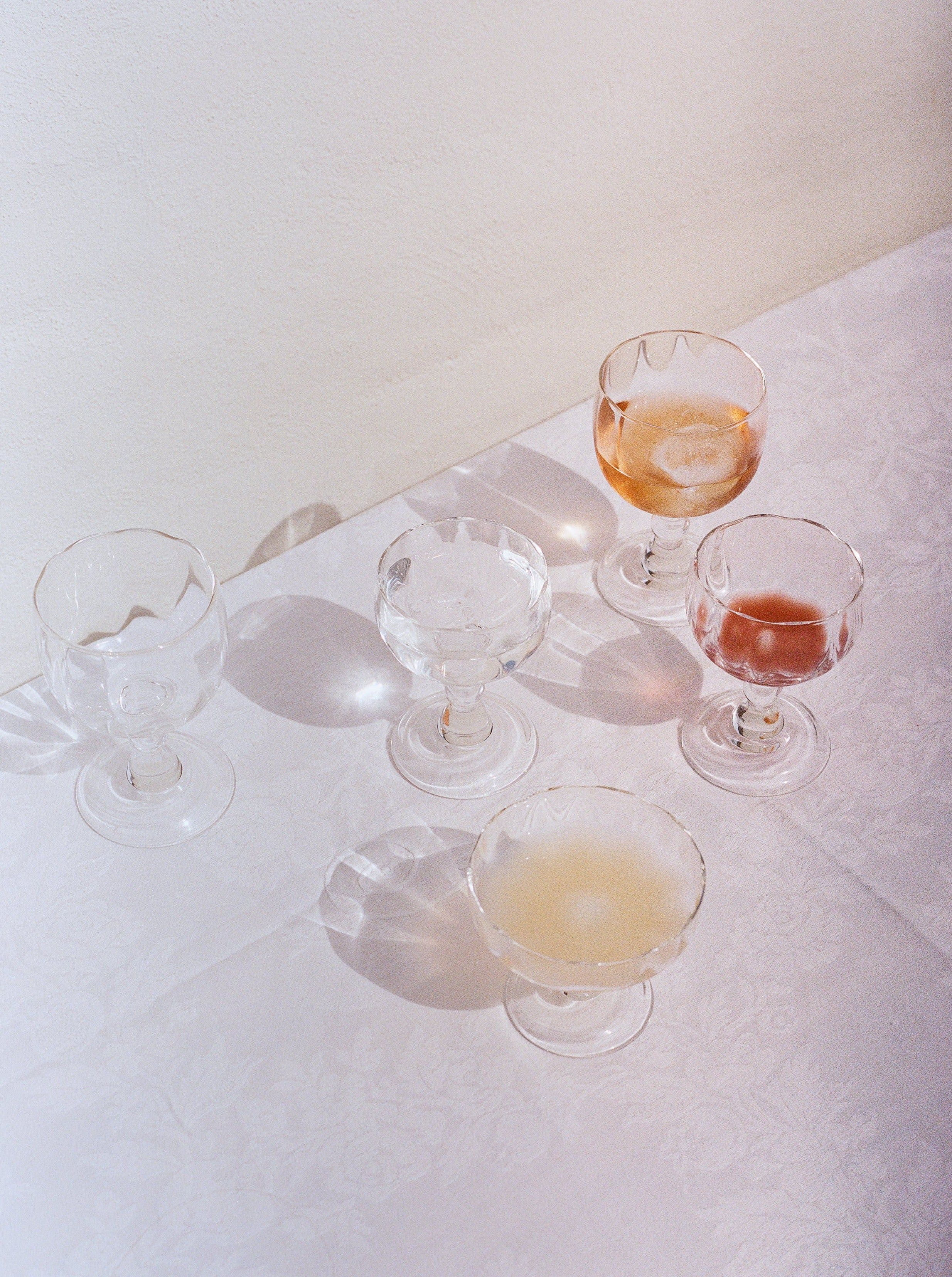 Five Alban Wine Glass Small goblets by Akua Objects with various amounts of liquid, arranged on a lace-covered table under soft lighting, creating delicate shadows and reflections.