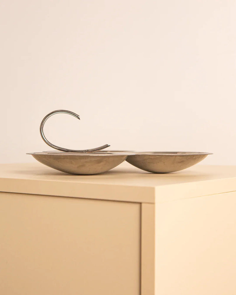 The "Cloverleaf" Shaped Vide-poches 80's by Treaptyque, a minimalist stainless steel sculpture with two shallow bowls and a curved design, is showcased on a light-colored angular pedestal against a plain light-colored wall.