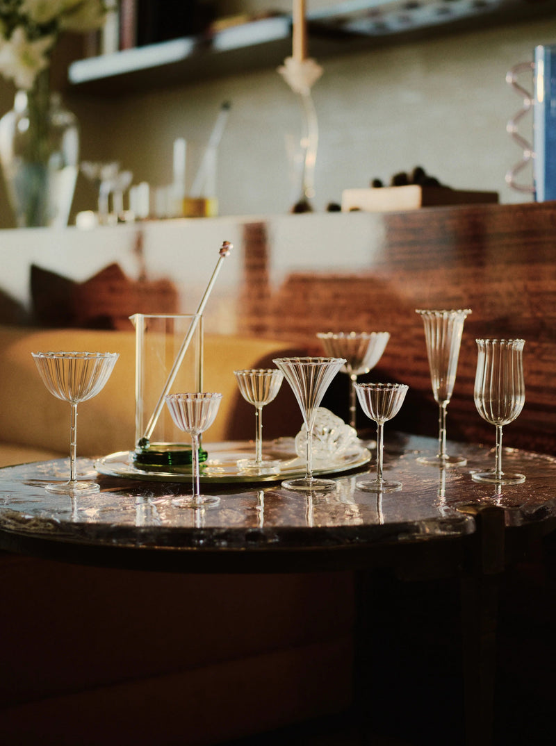 A set of 4 assorted, elegant empty glassware from the Angel Flute - Set of Four by Sophie Lou Jacobsen, including petite champagne flutes and wine glasses, arranged on a marble table. The background features a wooden shelf with various decor items and a vase with Angel Trumpet flowers, creating a sophisticated ambiance.