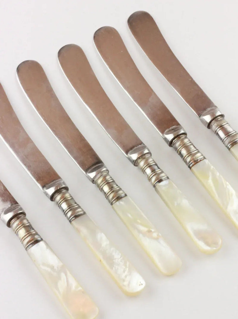 A set of six vintage flatware butter knives with stainless steel blades and iridescent mother-of-pearl handles, arranged in a row on a white background. Each Boga Avante Shop Mother of Pearl Knife features a decorative metal ring where the blade meets the handle, exuding timeless elegance.