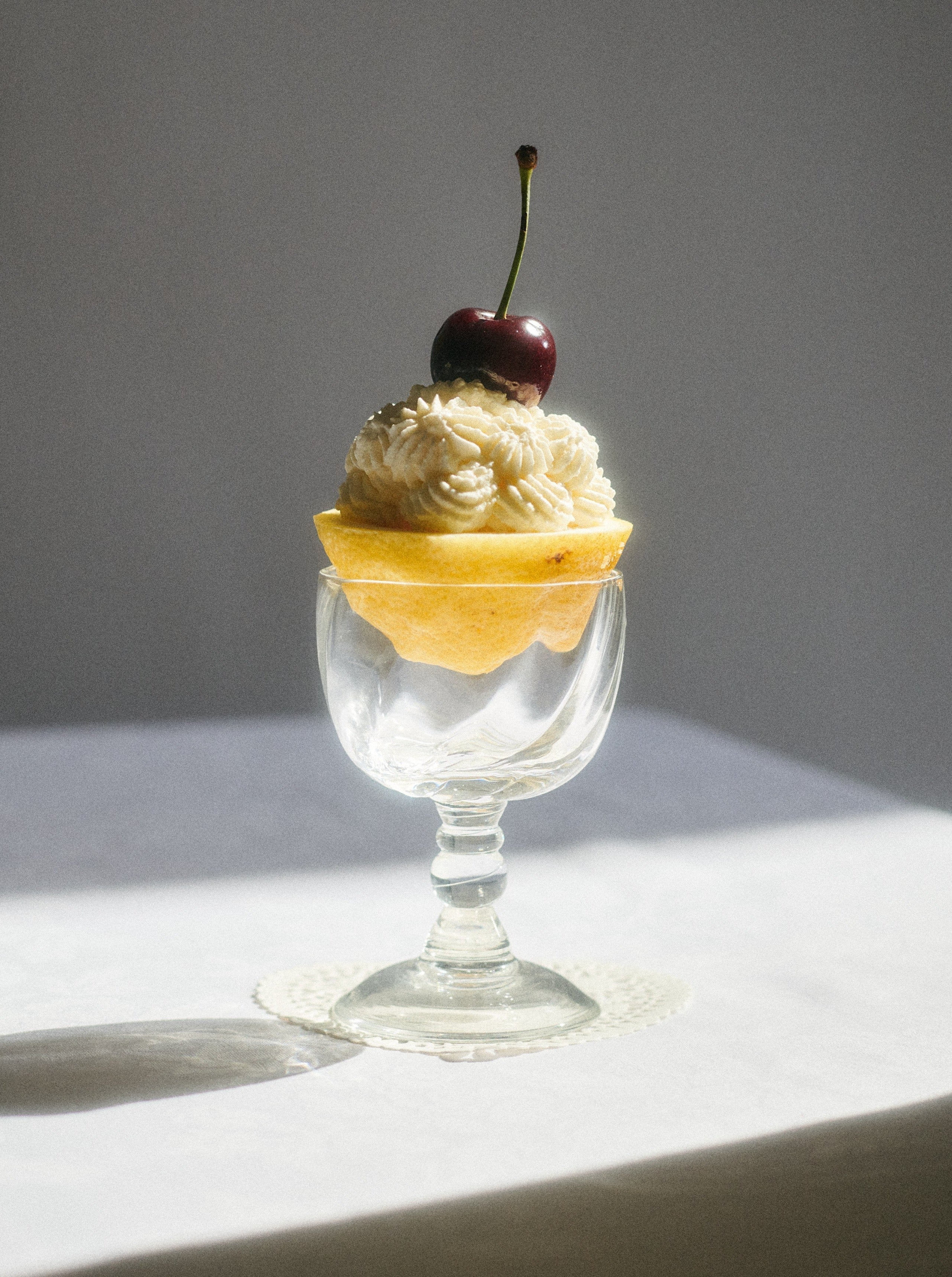 A delicate dessert in a clear Alban Wine Glass Small cup from Akua Objects, featuring layers of flaky pastry filled with a creamy, piped topping, crowned with a fresh cherry on top, all beautifully illuminated by natural lighting.