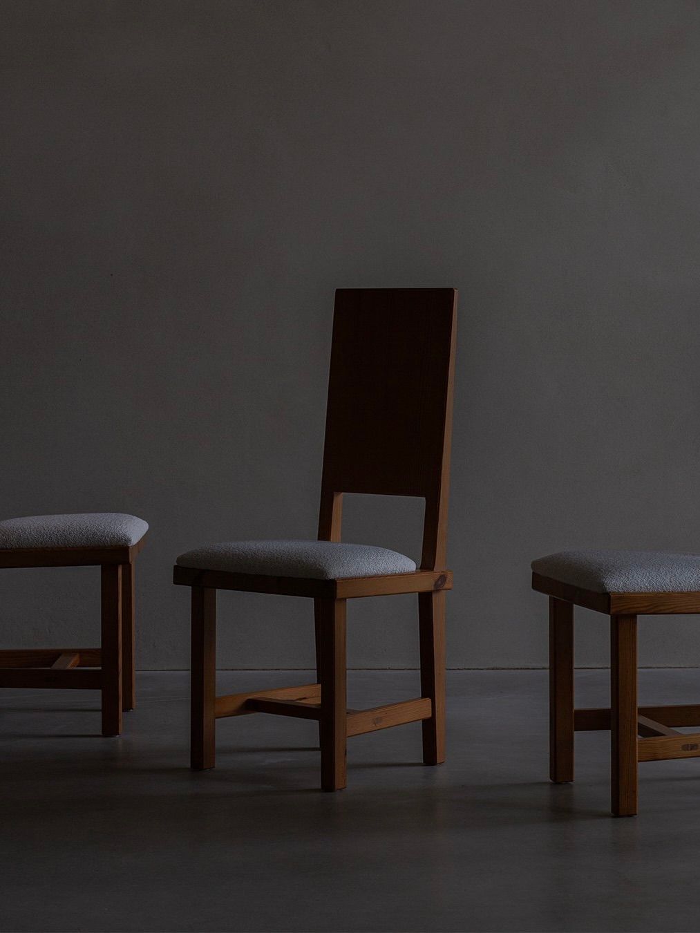 Three Bicci de' Medici Göran Malmvall chairs with gray cushions sit on a smooth floor against a dark wall. The central Model 553 chair has a taller backrest, while the side chairs have shorter ones. Showcasing Swedish design, the lighting is dim and moody.