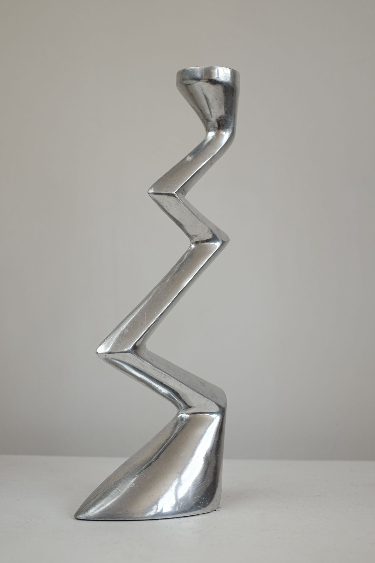 The Matthew Hilton ZigZag Candle Holder Large by septembre studios features a metallic silver finish and angular design, ideal for contemporary interiors and looks striking on a white surface against a light background.