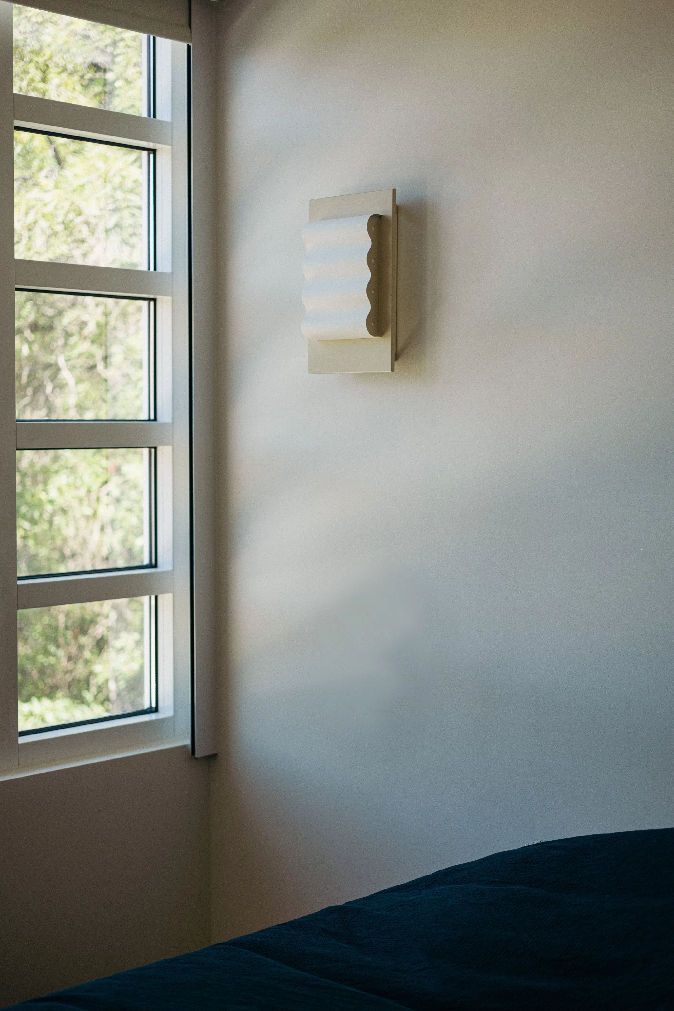 A minimalistic room with a light-colored wall features the elegantly handcrafted Violaine d'Harcourt Wall Light Frame OW S. Sunlight filters through a grid-patterned window, casting gentle shadows on a dark fabric, possibly from a bed or sofa, partially visible in the corner.