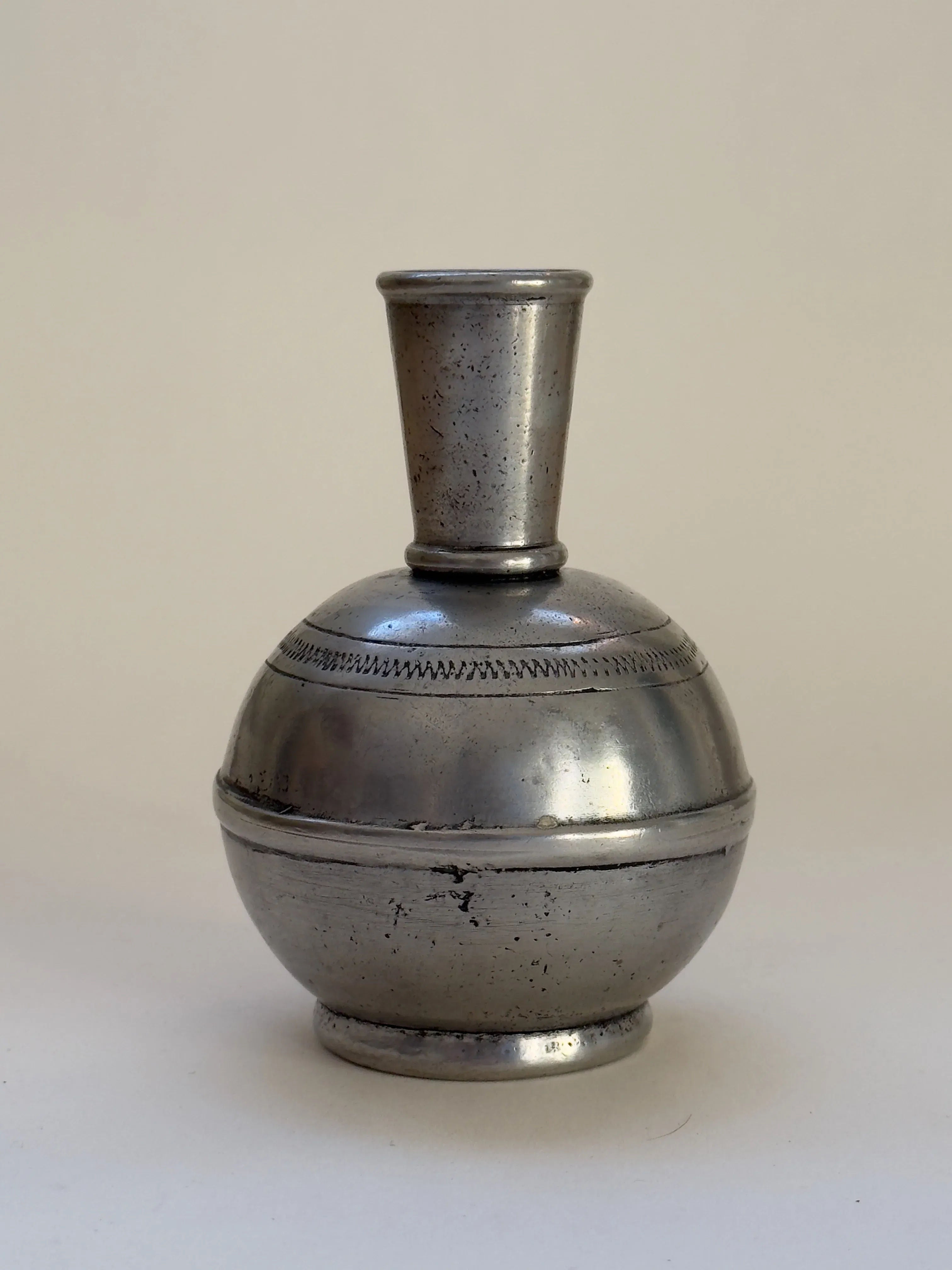 The Antique French Pewter Vase by Galerie Storm, from the 19th/20th Century, showcases a vintage design with a short, narrow neck and round body. It features engraved details enhancing its worn patina and stands on a small base with decorative etching against a plain background.