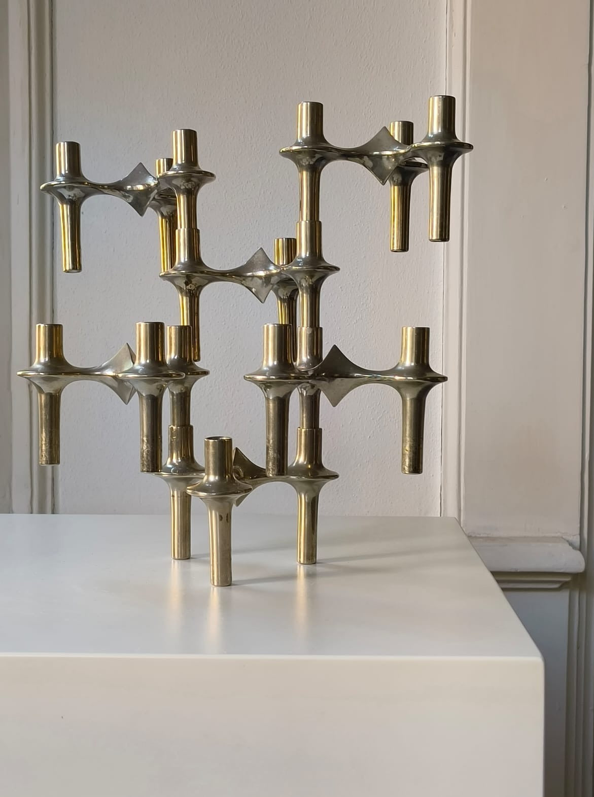 A Médecine Rare Franz Nagel Candlestick, with its sculptural and modular design made of shiny brass, is displayed on a white pedestal against a light-colored wall. The piece features multiple interconnected candle slots at varying heights, creating a geometric and contemporary aesthetic reminiscent of iconic Franz Nagel creations.