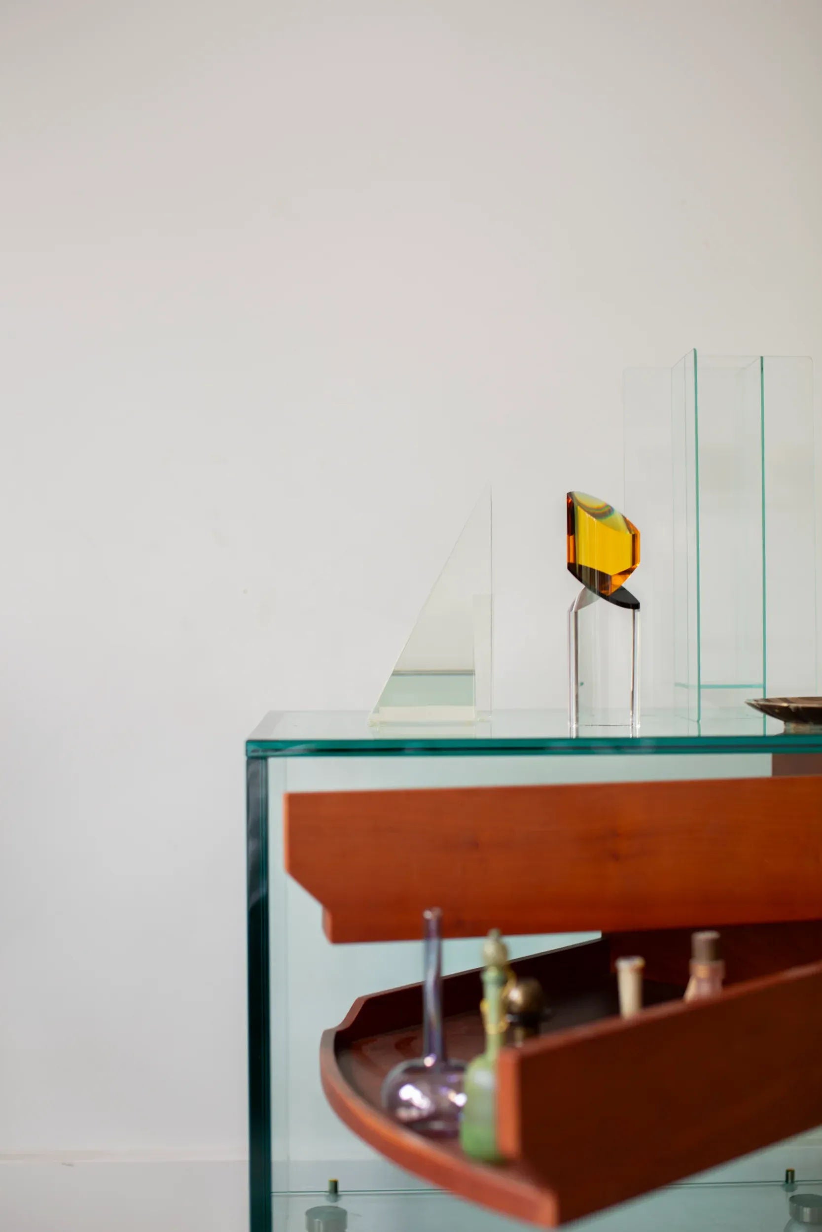 A modern minimalist scene featuring a tempered glass display table with geometric glass sculptures on top. In the foreground, a CU-RA-TED Tonelli Camicino Cabinet designed by Marco Gaudenzi has its wooden drawer partially open, revealing various small bottles and vials inside. The background is a plain white wall.