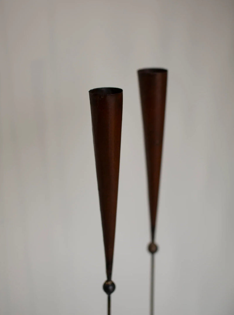 A close-up of two vertical, tapered wooden objects with a conical shape, resembling handmade candle holders or decorative spindles, set against a plain, light background. These 80's signed pieces by Pieter Spruyt have a dark brown finish and are minimalist in design from the CU-RA-TED brand appear.
