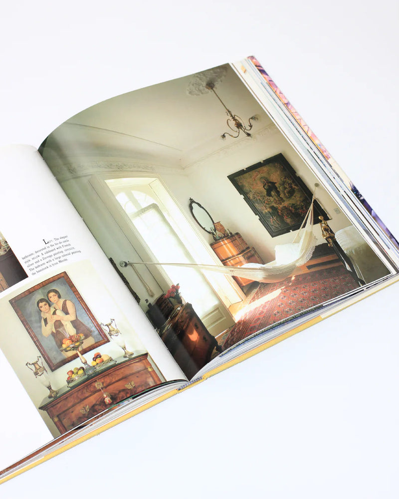 A "Casa Mexicana" book from Boga Avante Shop opens to reveal an elegantly designed interior with a hammock, vintage decor featuring a wooden dresser, ornate mirror, framed paintings, and a patterned rug—all embodying the charm of domestic architecture.
