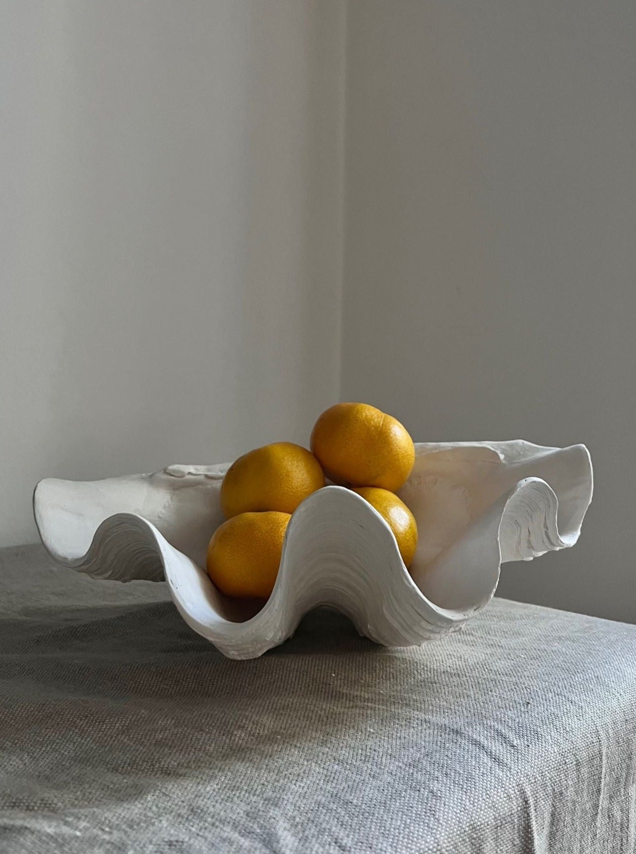 The Médecine Clam Shell Bowl - Handmade Plaster Sculpture, in white, holds four yellow lemons and sits on a textured gray fabric surface against a softly lit, plain wall.