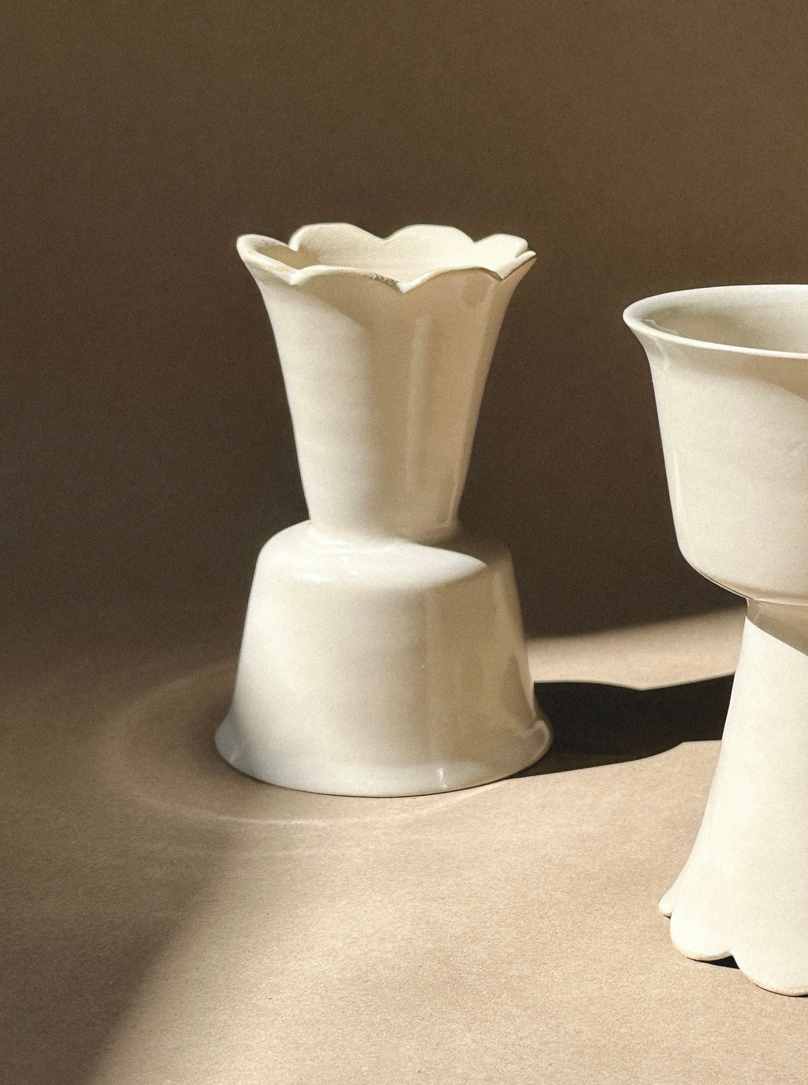 A minimalist, beige-toned image features two white Flowery Wine Goblets by noki ceramics with scalloped edges. The handmade stoneware pieces are set against a neutral background, with soft shadows enhancing their shapes and textures. The arrangement emphasizes their simple yet elegant design.