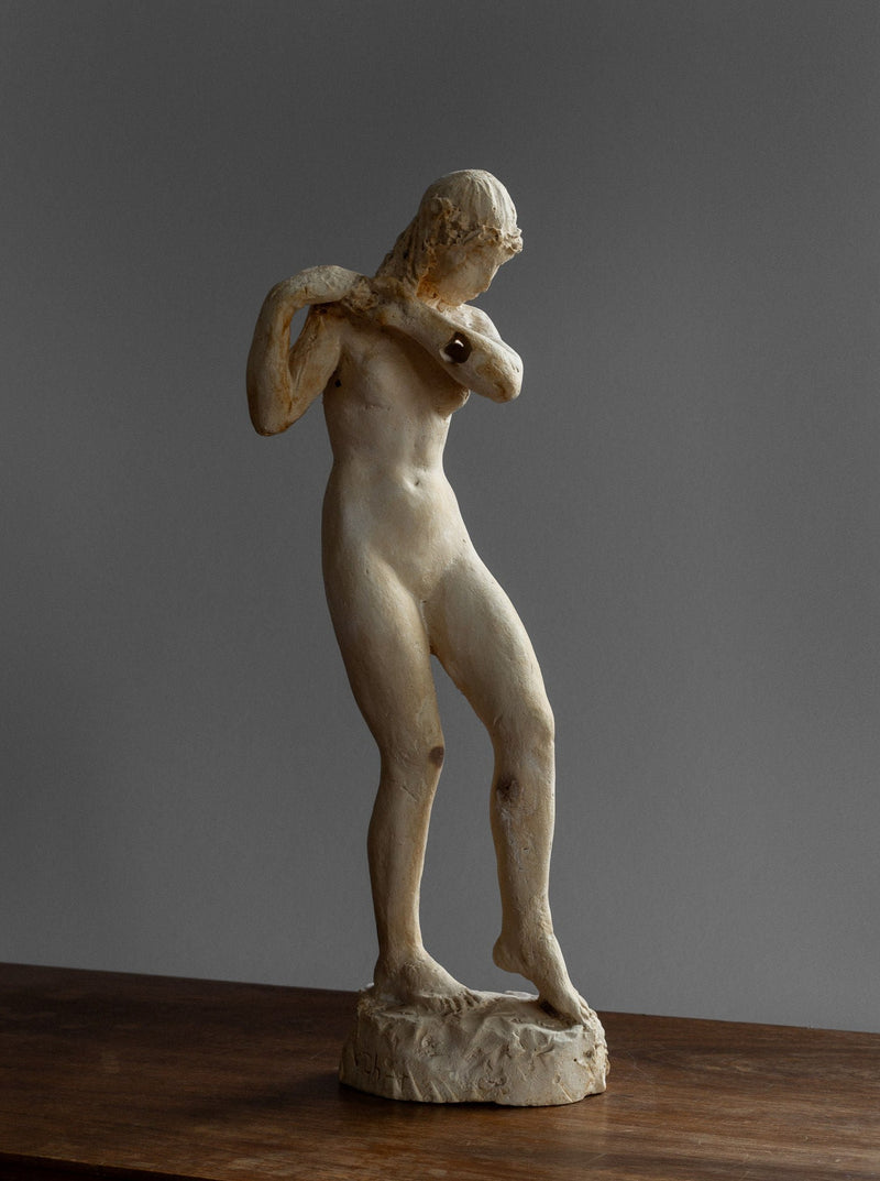 A weathered, classical-looking plaster sculpture of a nude figure, named the Vintage Neoclassical Sculpture from Spigel, stands on a wooden surface. Thought to be of Swedish origin from the 1920s-30s, the figure is poised with an arm resting against its chest and the other at its side, creating a graceful, slightly twisted pose. The background is plain and gray.