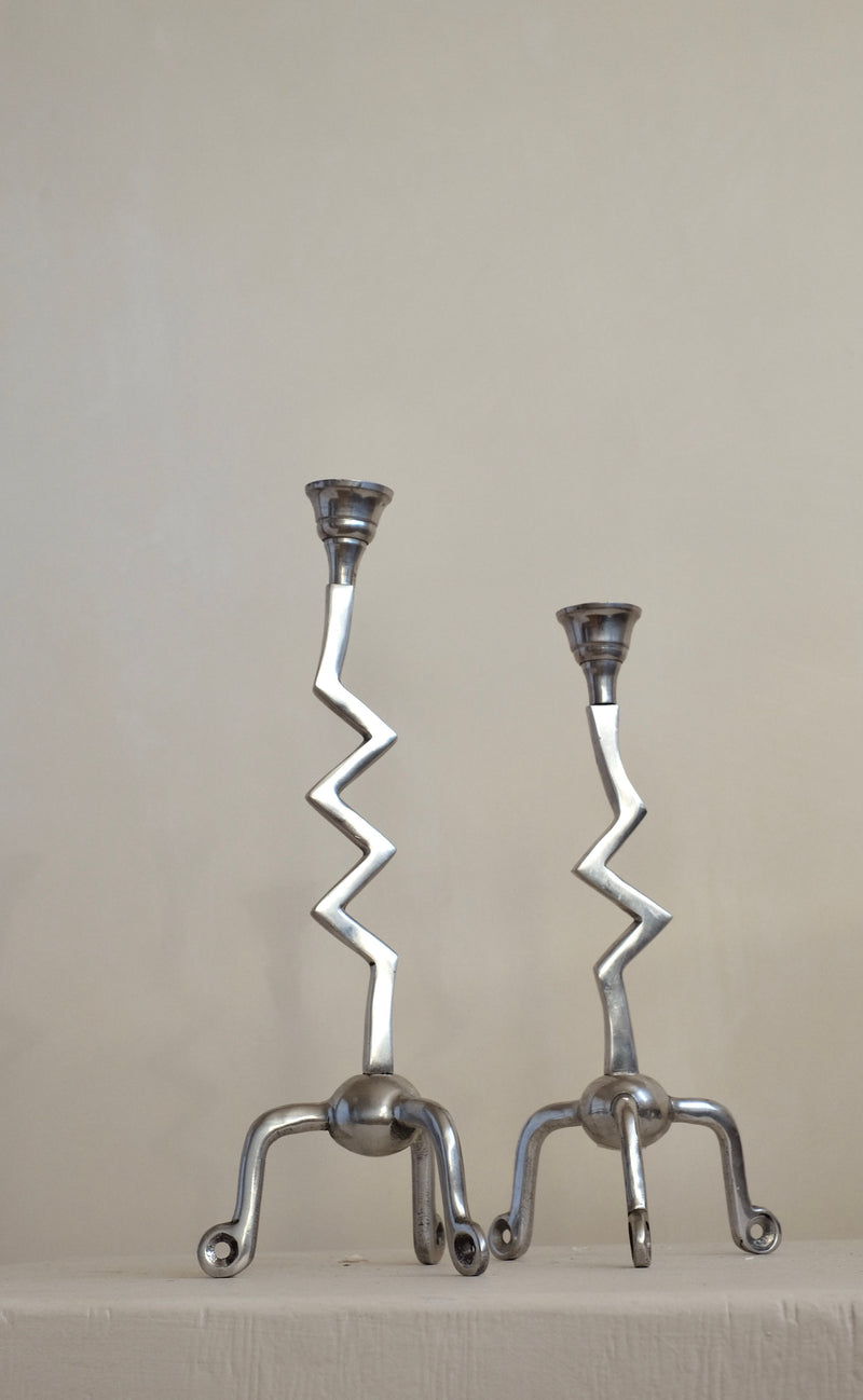 A pair of Zigzag Candleholders from septembre studios, featuring zigzag stems and tripod bases, stand side by side against a plain background. Their distinctive design brings an artistic flair to the setting, making them a genuine zilver treasure.