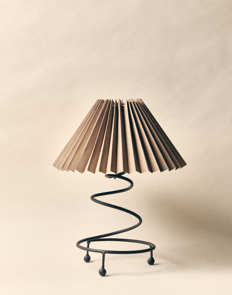 The SÒLER 'Como' Table Lamp features a pleated beige shade and a black wrought-iron spiral base, beautifully set against a matching beige background.
