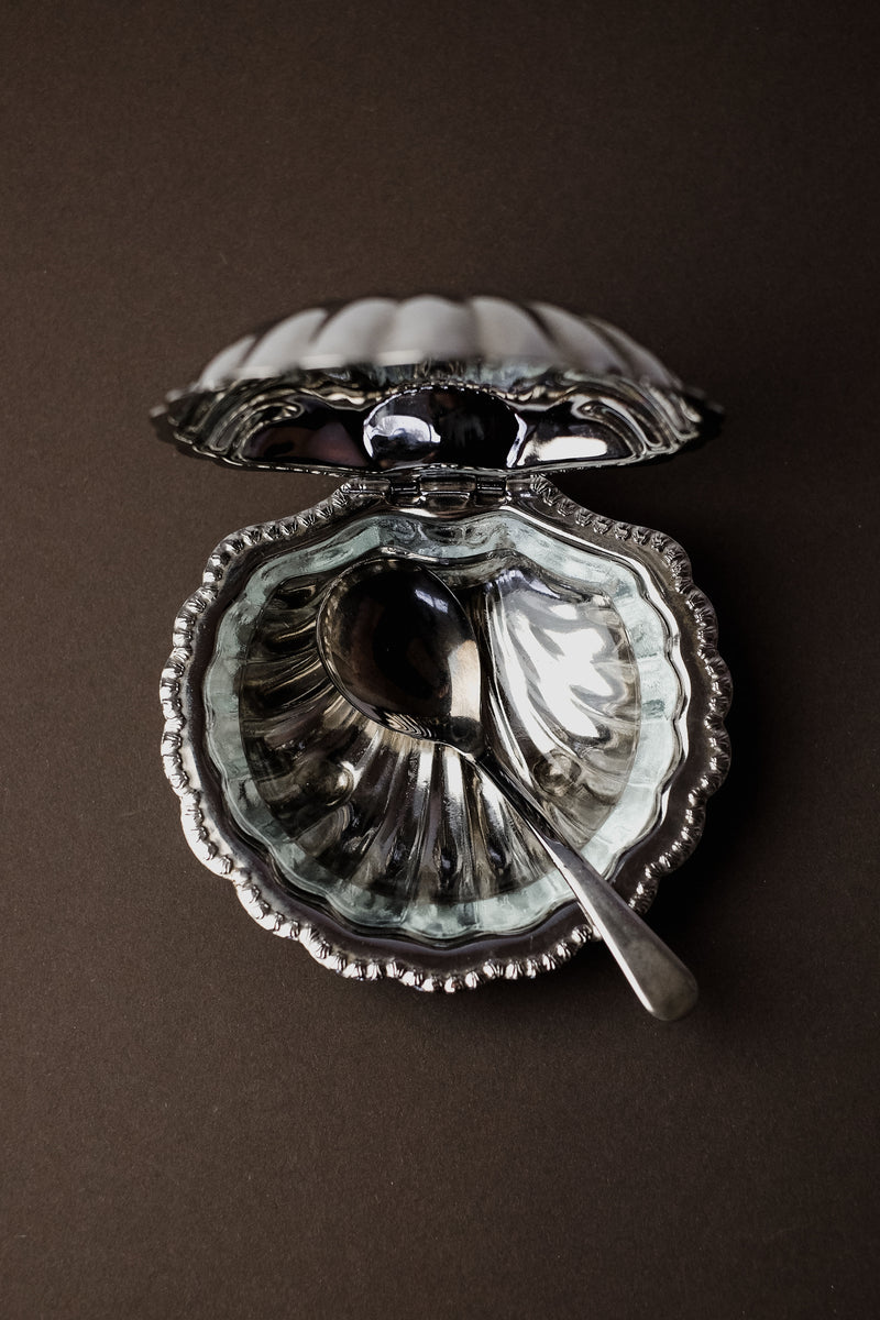 The Shell Shaped Butter Dish with Spoon by septembre studios features a scalloped seashell design that opens to reveal a shiny interior, complemented by a small silver spoon. The partially visible lid's reflective finish adds an elegant charm to your dining experience.