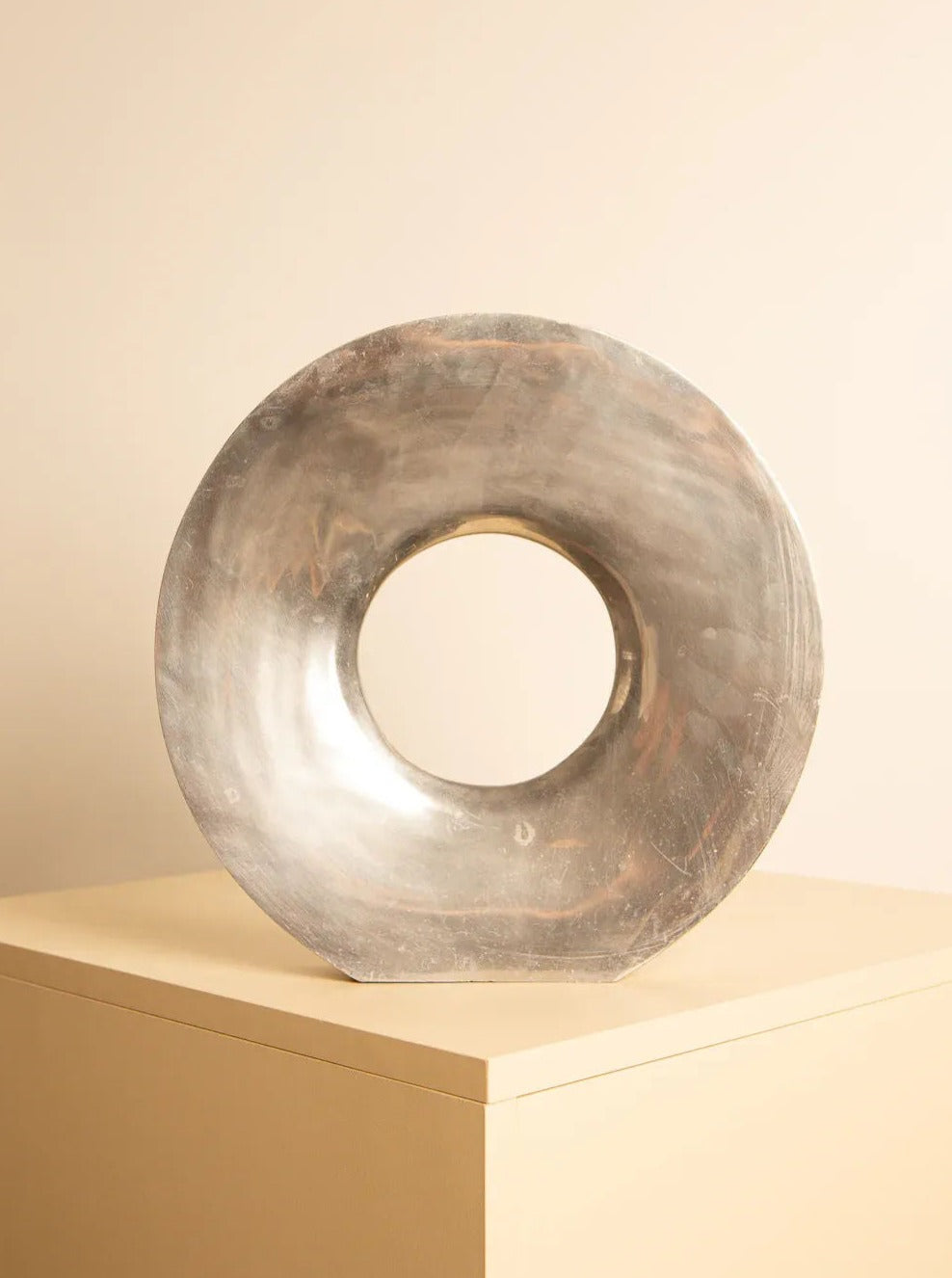 A circular, metallic sculpture with a large central hole, resembling a contemporary doughnut shape, is displayed on a beige pedestal against a neutral background. The surface has a reflective, slightly weathered finish, showcasing exquisite Italian craftsmanship. This piece is the "Donut" Vase in Aluminium 80's by Treaptyque.