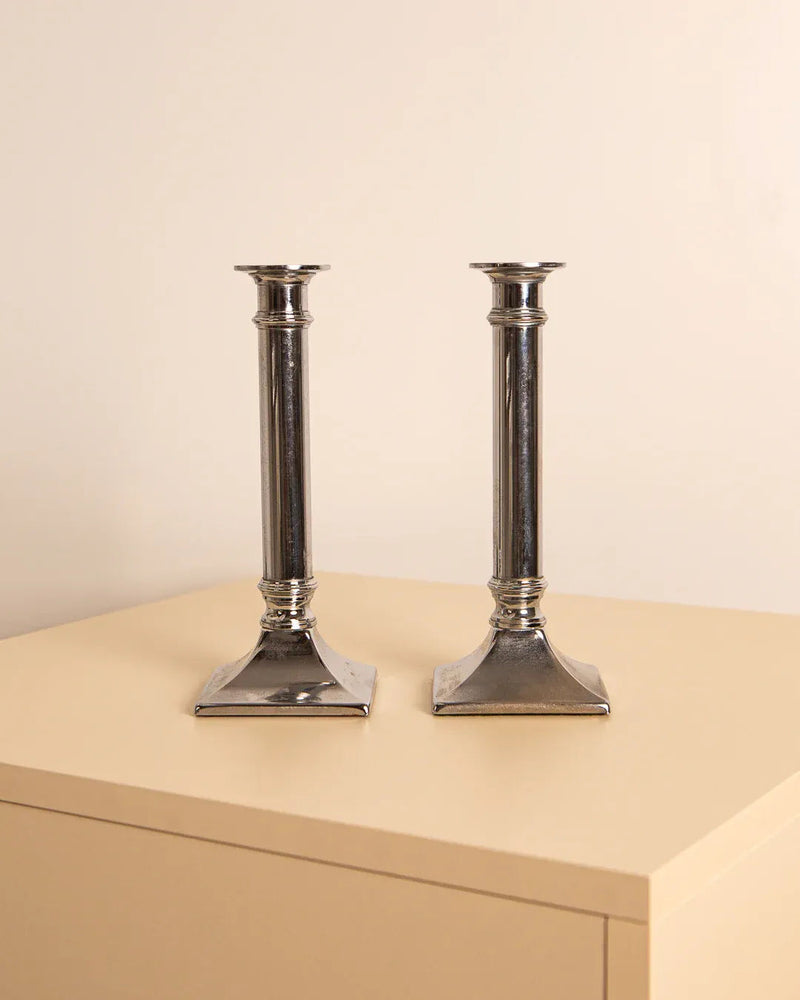 A pair of "Colonne" metal candleholders from the 80s brand Treaptyque is elegantly displayed on a light beige wooden table against a plain cream wall, adding a vintage industrial aesthetic.