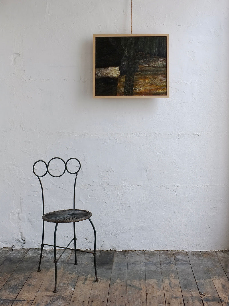 A minimalist room showcases a rustic wood floor and metal chair with a circular backrest. A Veter Vintage "Vintage Landscape Painting, Oil on Canvas," featuring earthy tones, is mounted on the white textured wall in a light wood frame.