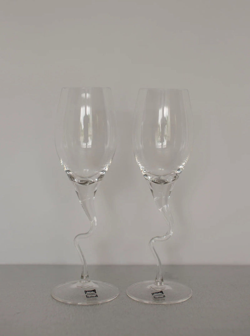 Two clear, intricately designed Set of Vintage Handmade Swirl Champagne Flutes by CU-RA-TED with twisted stems are placed side by side on a gray surface. The artisan-made, artistic shape of the stems adds a vintage charm and sophisticated touch to the otherwise classic glassware.