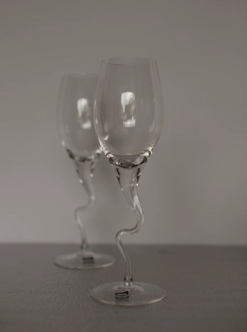 Two empty, clear champagne flutes with elegantly twisted stems are placed against a plain background. The artisan-made design of the Set of Vintage Handmade Swirl Champagne Flutes from CU-RA-TED adds a touch of vintage charm to the simple glassware. The lighting is soft, casting subtle reflections on the glass surfaces.