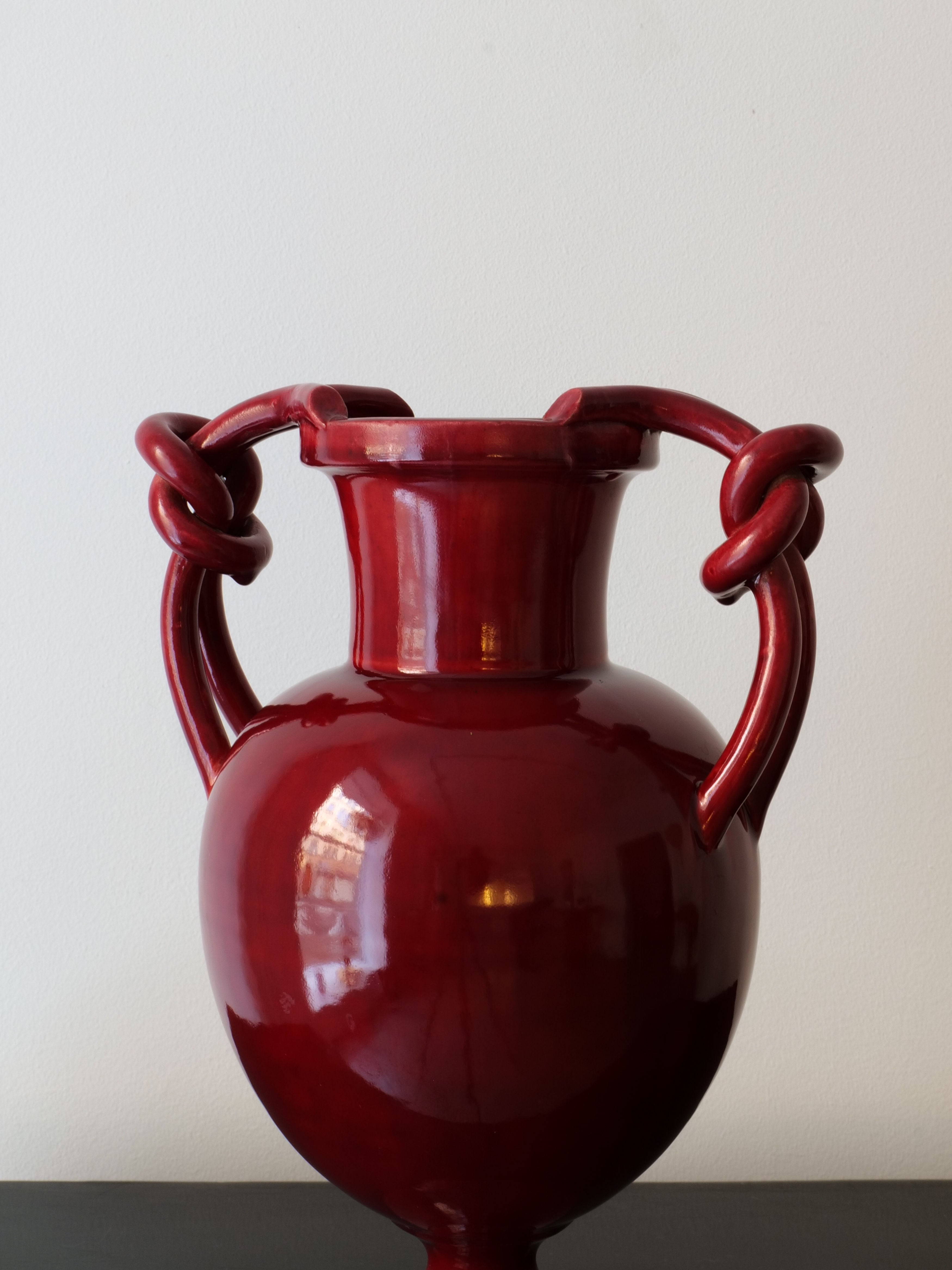 The Baluster Vase Jérôme Massier by Collection apart, in a glossy, deep red ceramic reminiscent of French Art Nouveau, features two decorative handles. It sits elegantly on a black surface against a plain white background. The design echoes the style of Vallauris ceramics, with a rounded body and wide neck accompanied by gracefully looping handles.