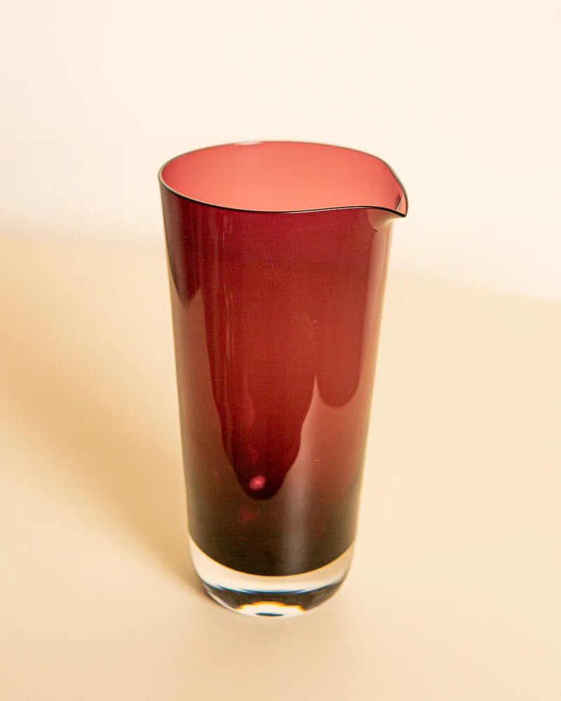 A tall Treaptyque Bordeaux Murano Glass Pitcher 80's in deep maroon with a spout is set against a neutral backdrop. It showcases the glossy texture and transparent base typical of Italian craftsmanship, epitomizing artisanal excellence.