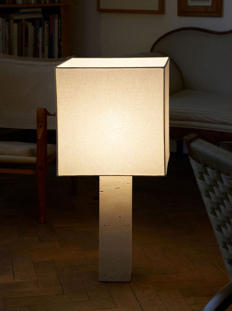 A modern, minimalist floor lamp with a beige square shade and a rectangular base glows softly in a cozy living room with wooden floors. The Nassi Lamps' Roma I Squared White Travertine casts a gentle light that complements the space beautifully. In the background, there is a beige armchair, a wooden bookshelf filled with books, and a beige couch, all embodying the essence of modernism.