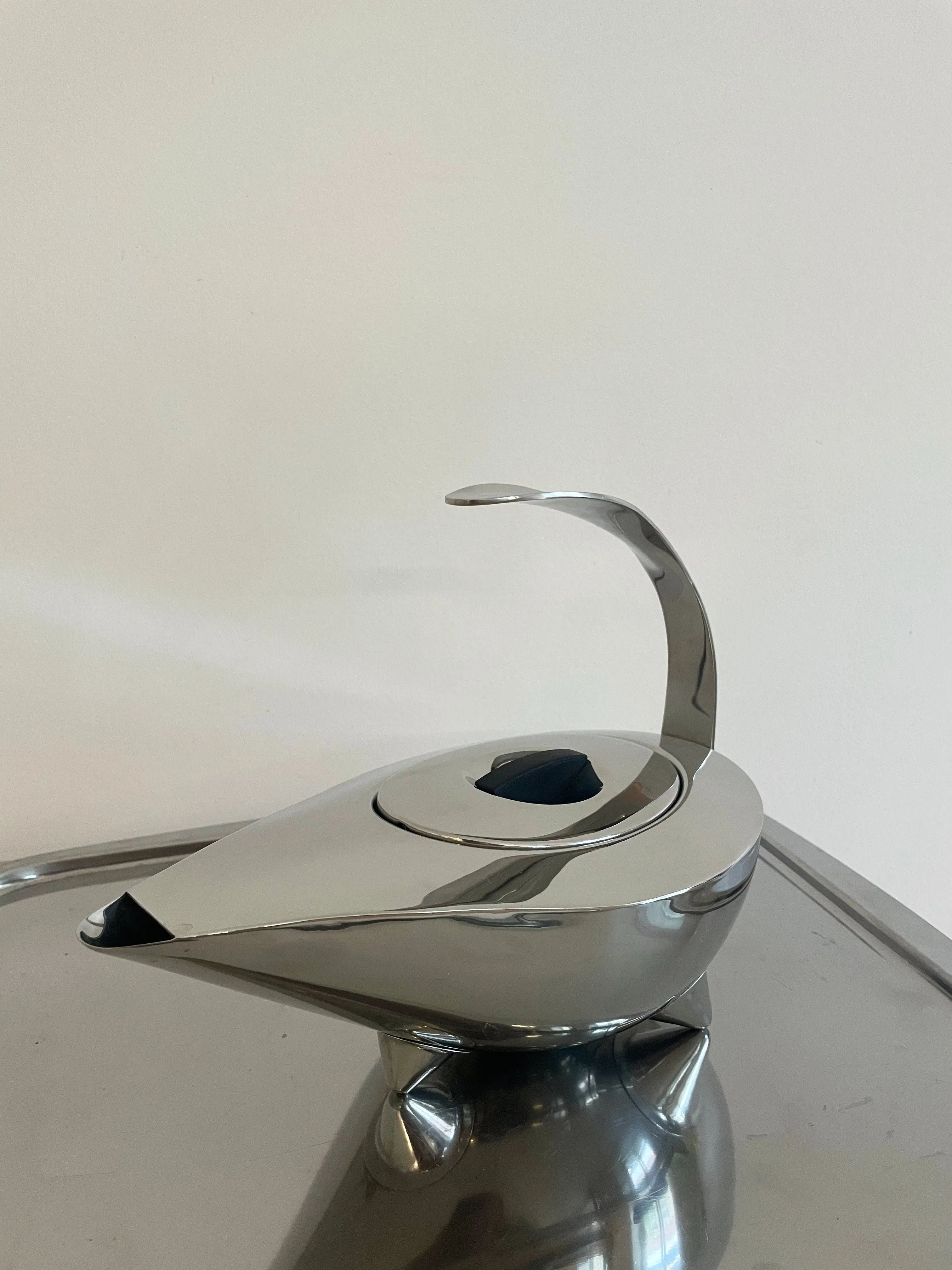 A modern Collect Cph 1980 Bodum Naoko teapot, crafted from stainless steel with a sleek, curved handle and streamlined design, sitting on a reflective surface against a plain white background.