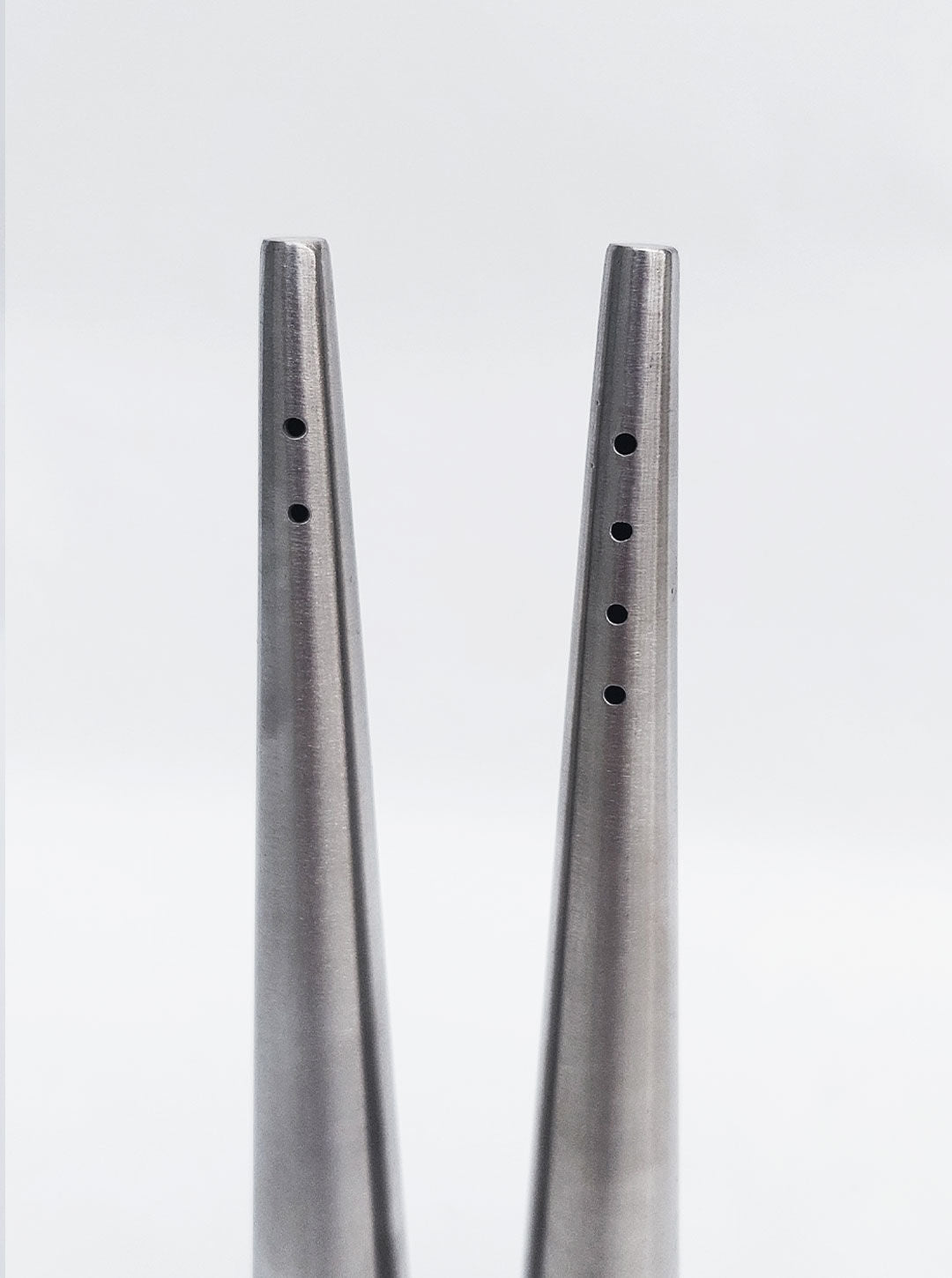 Close-up of the Conic Pepper and Salt Set, two luxurious mate steel, silver metallic conical objects with small circular holes aligned vertically near their tips. The objects are vertically oriented with the tips pointing upwards against a plain white background, perfect for an exquisite dining experience. From Les Objoies.