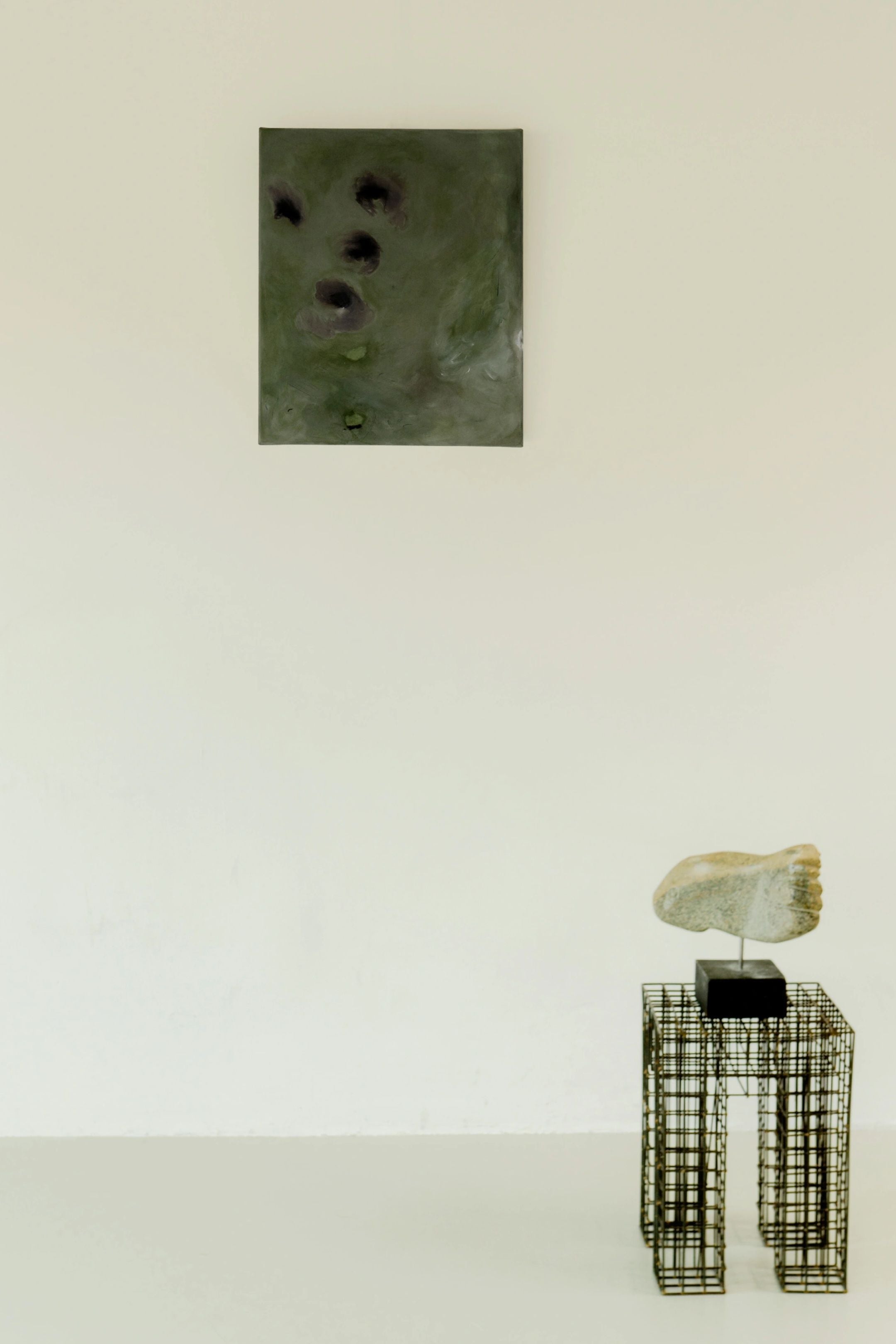 A minimalist art display features an original piece: "Let go of gravity," a dark abstract painting by Yeliza Gevorgyan on a white wall. It is complemented by a stone foot sculpture on a small black pedestal, with the unruly forms elegantly poised on the wireframe stand, inviting contemplation and intrigue.