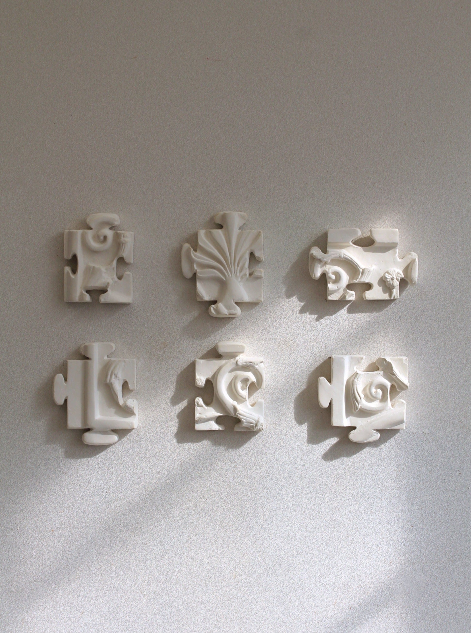 Six intricately carved white plaster pieces, reminiscent of jigsaw puzzle segments, are arranged in a striking wall sculpture of two vertical columns against a plain light-gray background. This sexiptyque features unique patterns and designs, casting gentle shadows on the wall. The artwork is named "Puzzle Antique" and is crafted by the renowned brand Marina Mankarios.