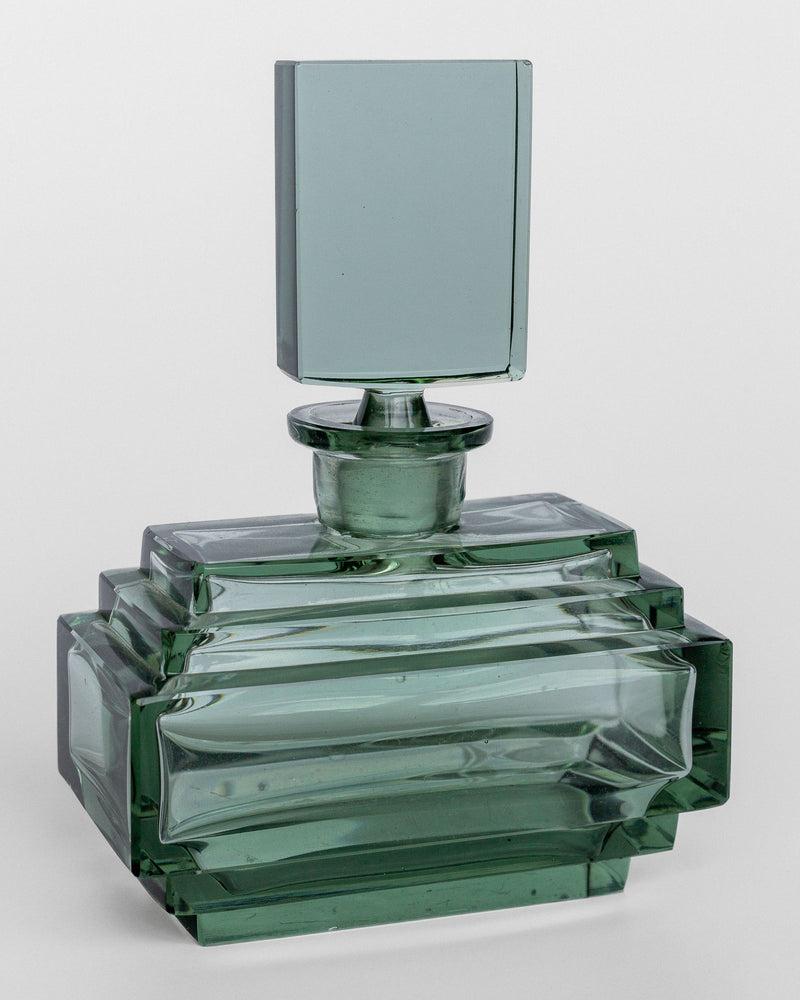The Spigel Art Deco Perfume Bottle, inspired by the 1930s, showcases a clear, geometrically-styled design with a square stopper. The intricately multi-layered rectangular pattern highlights modern elegance and Czech craftsmanship; the transparent glass subtly hints at a green tint.