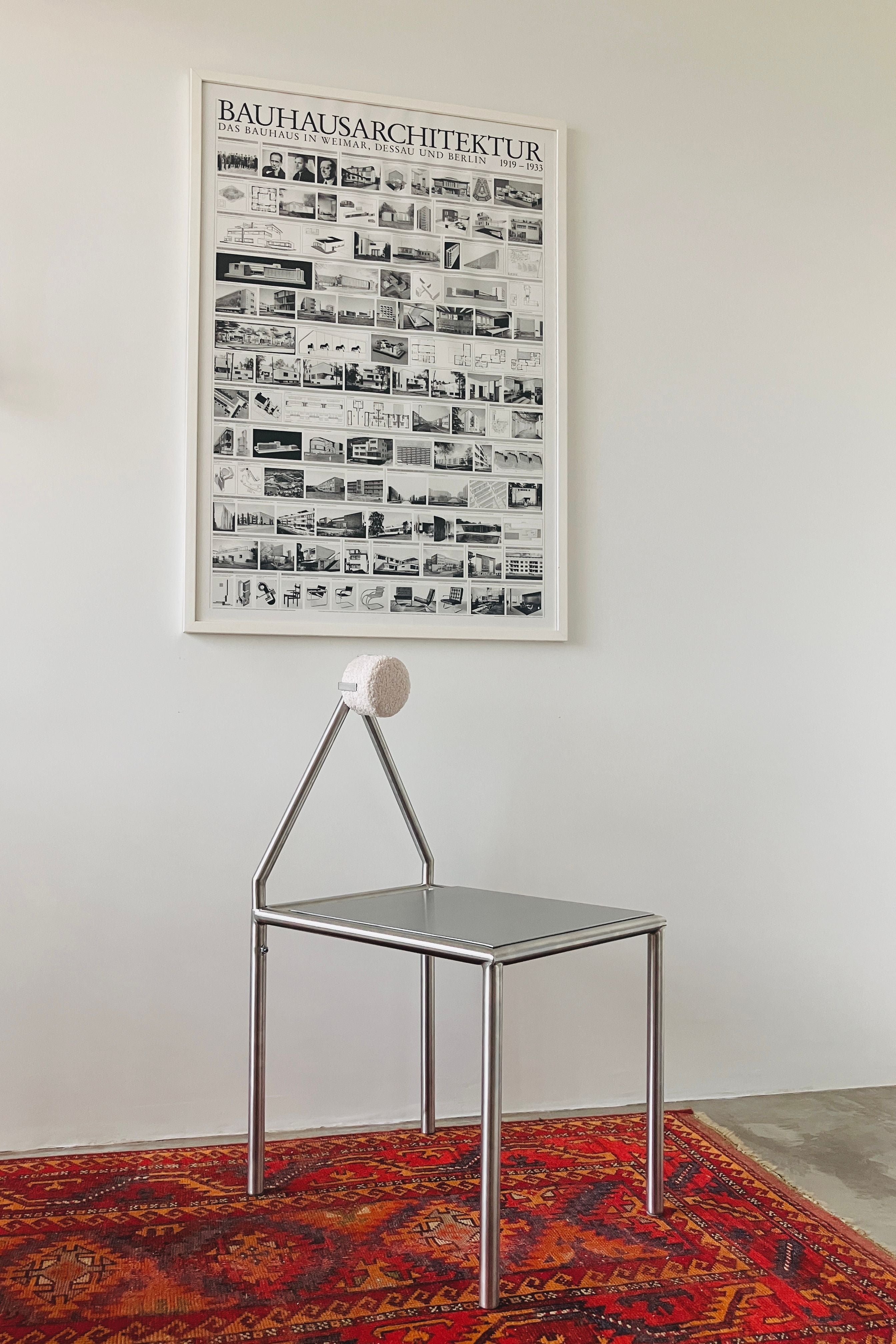 An 'H01' Stainless Steel Chair by HT. with a modern geometric design sits on a red patterned rug beneath a framed "Bauhaus Architektur" poster featuring black-and-white architectural images on a white wall.