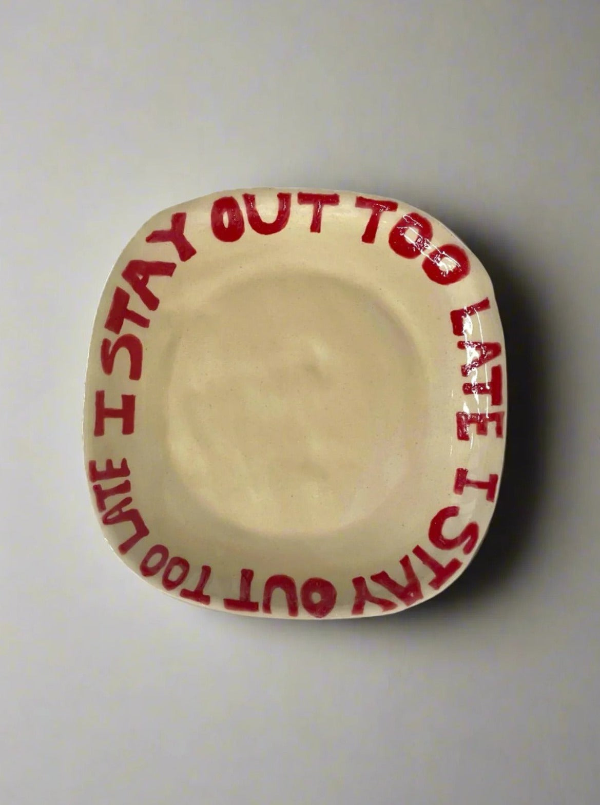 The "I Stay Out Too Late Tray in Red" from L&Clay Ceramics is a custom-made, square-shaped ceramic plate with rounded corners. It features the bold red text "I STAY OUT TOO LATE" around its rim and is showcased against a plain light-colored background.