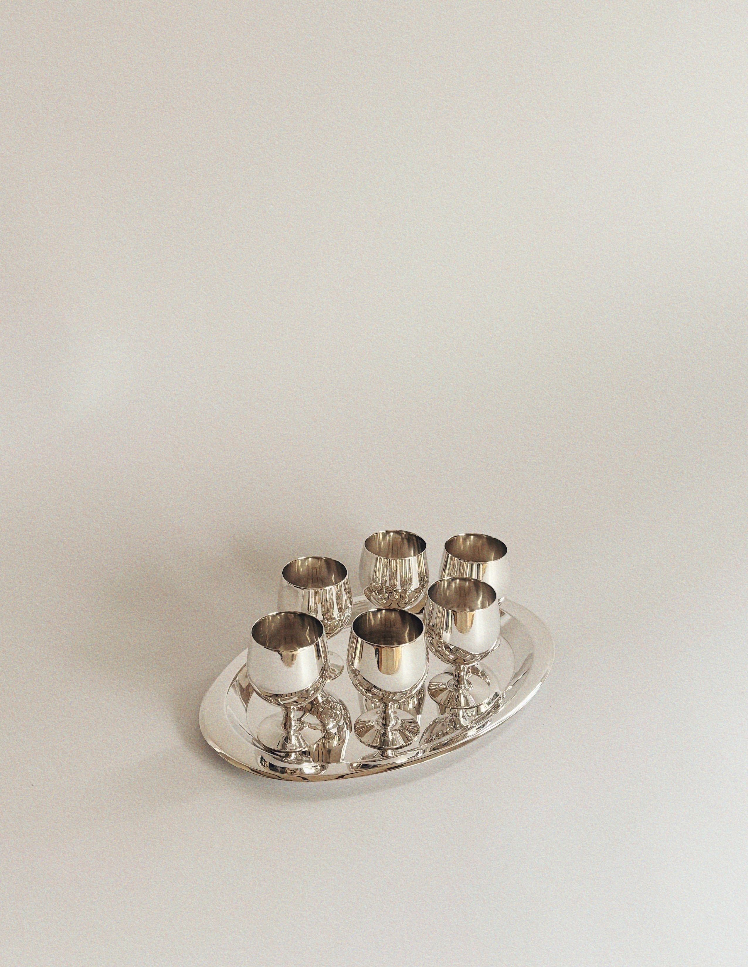 A rare Silver Plated 70's Liquor Server from Huerto, featuring six silver-plated liquor glasses, is elegantly arranged on a matching oval tray against a plain white background. The reflective surfaces of the goblets and tray evoke the polished sophistication reminiscent of communist-era Poland.