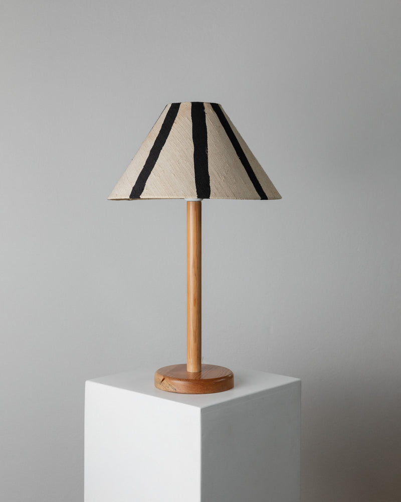 The Bottega Jacobs Table Lamp by Solbackens Svarveri showcases a wooden base and cylindrical stand on a white pedestal. It has a hand-painted linen shade with beige and black vertical stripes, reflecting Swedish mid-century design against a light gray wall.