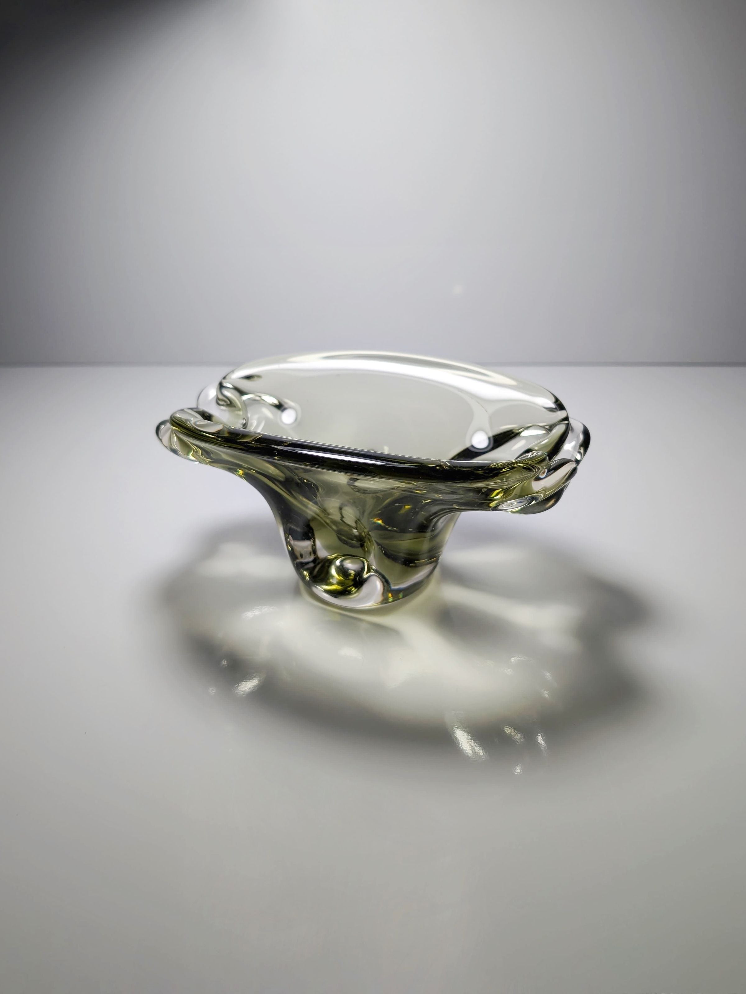 The Bohemian Smoked Glass Ashtray by Jan Beránek from the 1960s, available at Dodo Vintage, is artistically shaped with fluid curves and reflections, reminiscent of classic design. It sits elegantly on a smooth white surface under soft lighting that casts gentle shadows.