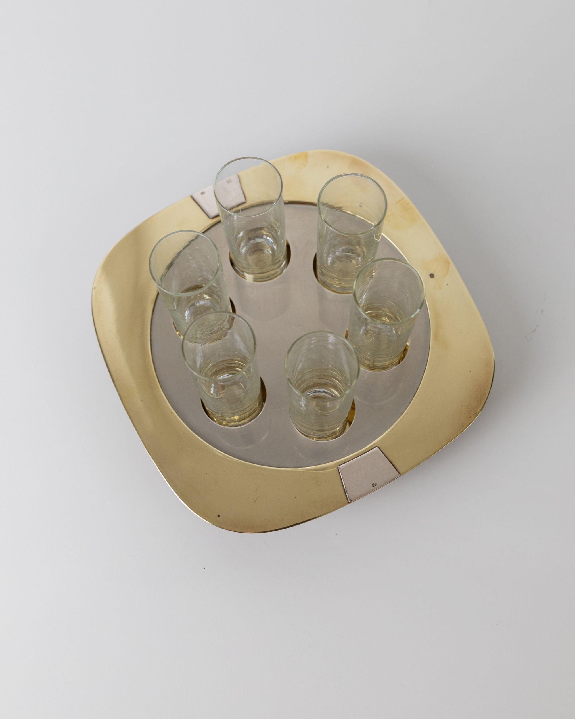 The "Set Shot Glasses and Tray - Mid Century Design 70s" by Bottega Jacobs features a square gold tray with smooth, rounded edges, holding six vintage clear shot glasses in a circular arrangement. Slight discoloration and scratches enhance its mid-century charm against a plain white background.