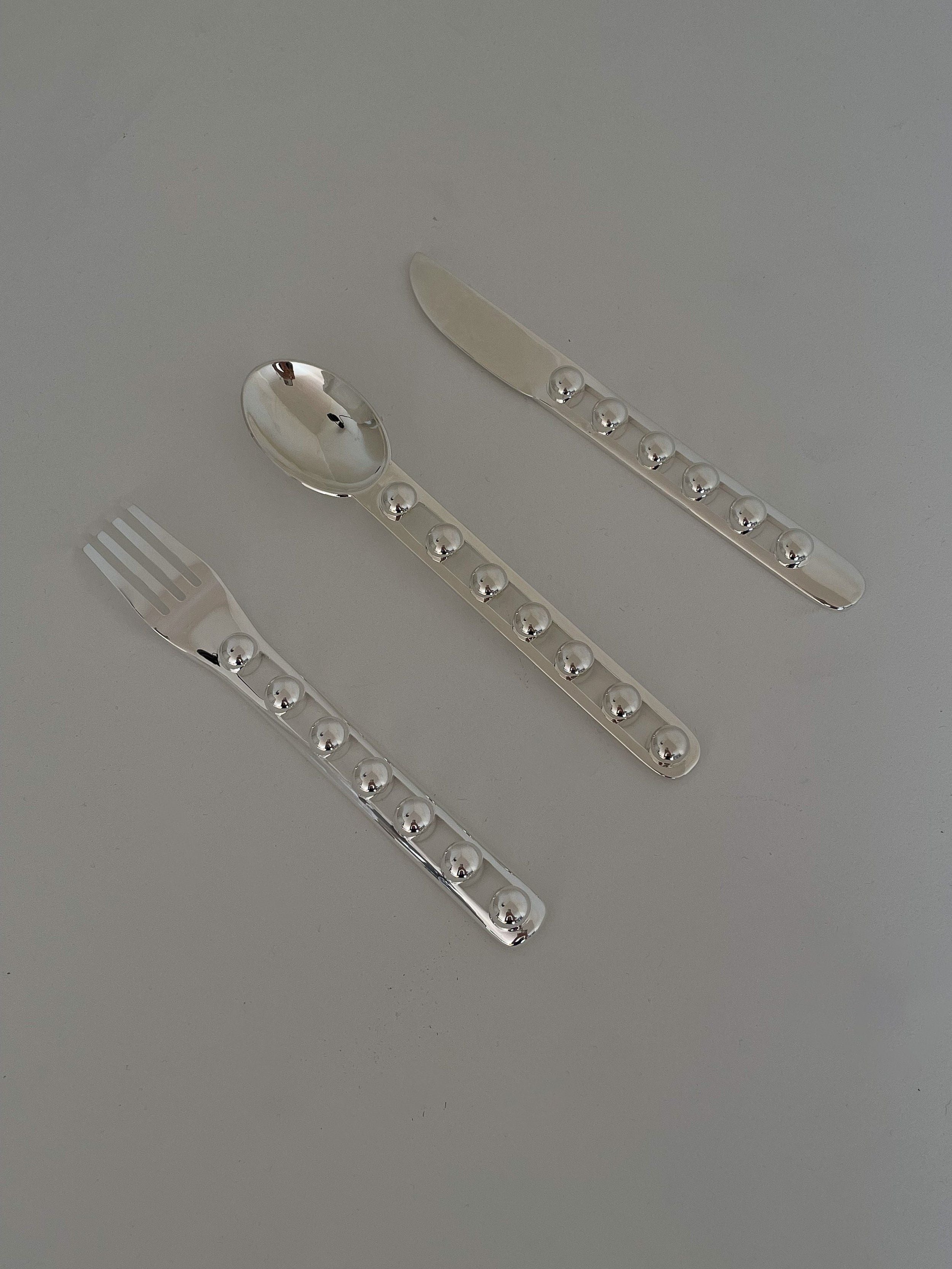 The "Seven Ball" Cutlery Set by Jessi Burch is displayed on a gray background, highlighting its functional art that includes a fork, spoon, and knife. Each piece boasts uniquely textured handles decorated with raised circular designs.