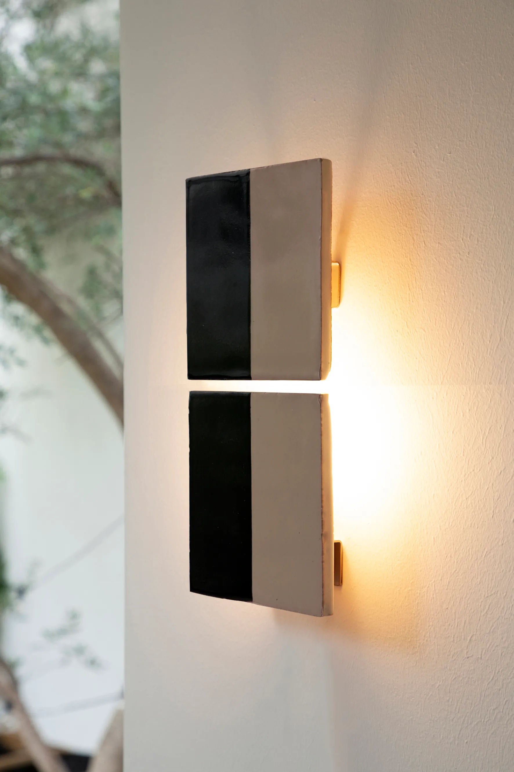 The Tiles Line N by Violaine d'Harcourt is a modern ceramic wall sconce featuring black and beige rectangular panels. Expertly crafted, it casts a warm, soft glow on a light-colored wall, illuminating a tree with green leaves in the background.
