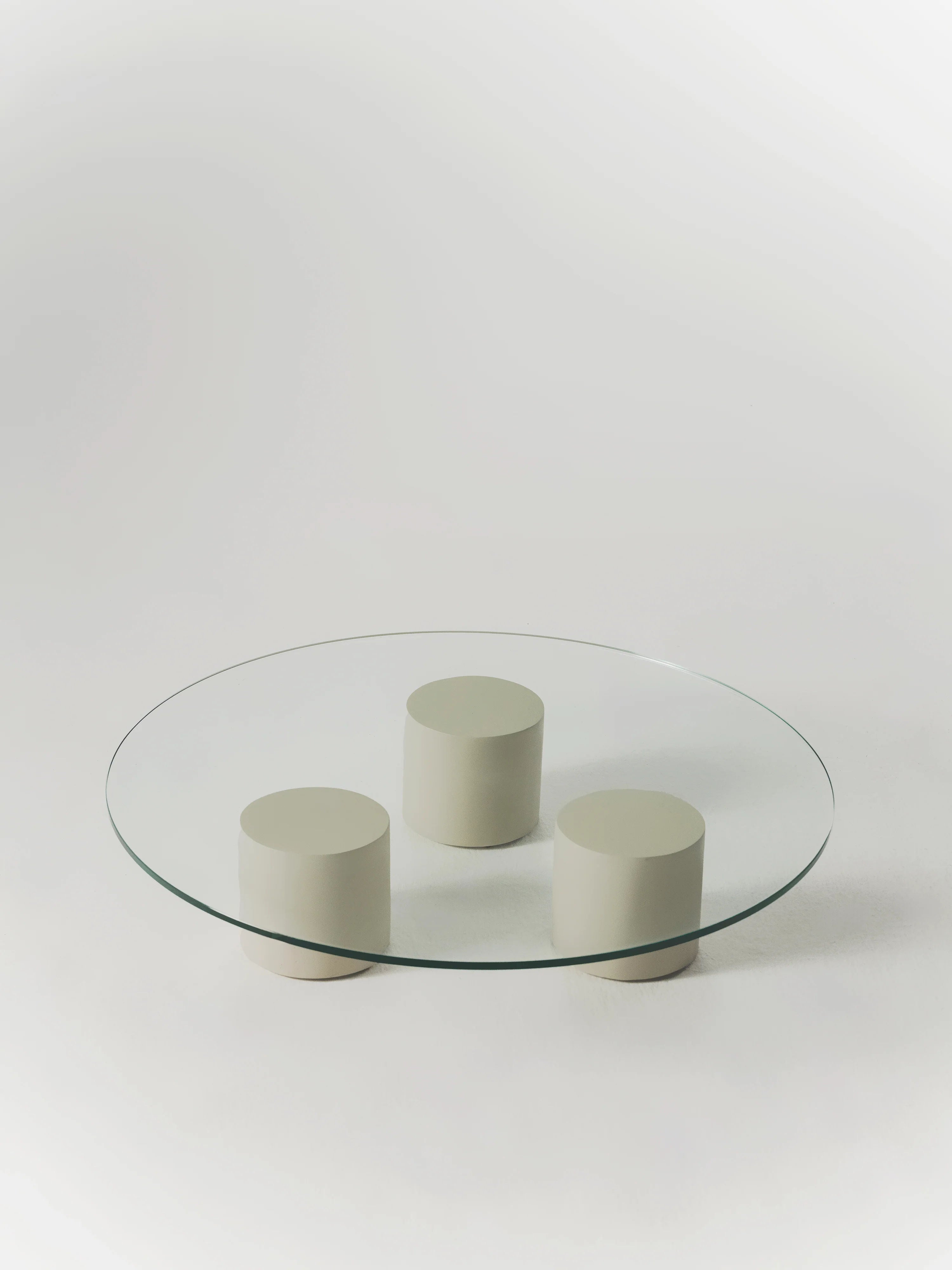 The Carmene Glass Coffee Table by MarlotBaus displays a round tempered glass top with three cylindrical beige supports in a triangular arrangement on a light grey backdrop.