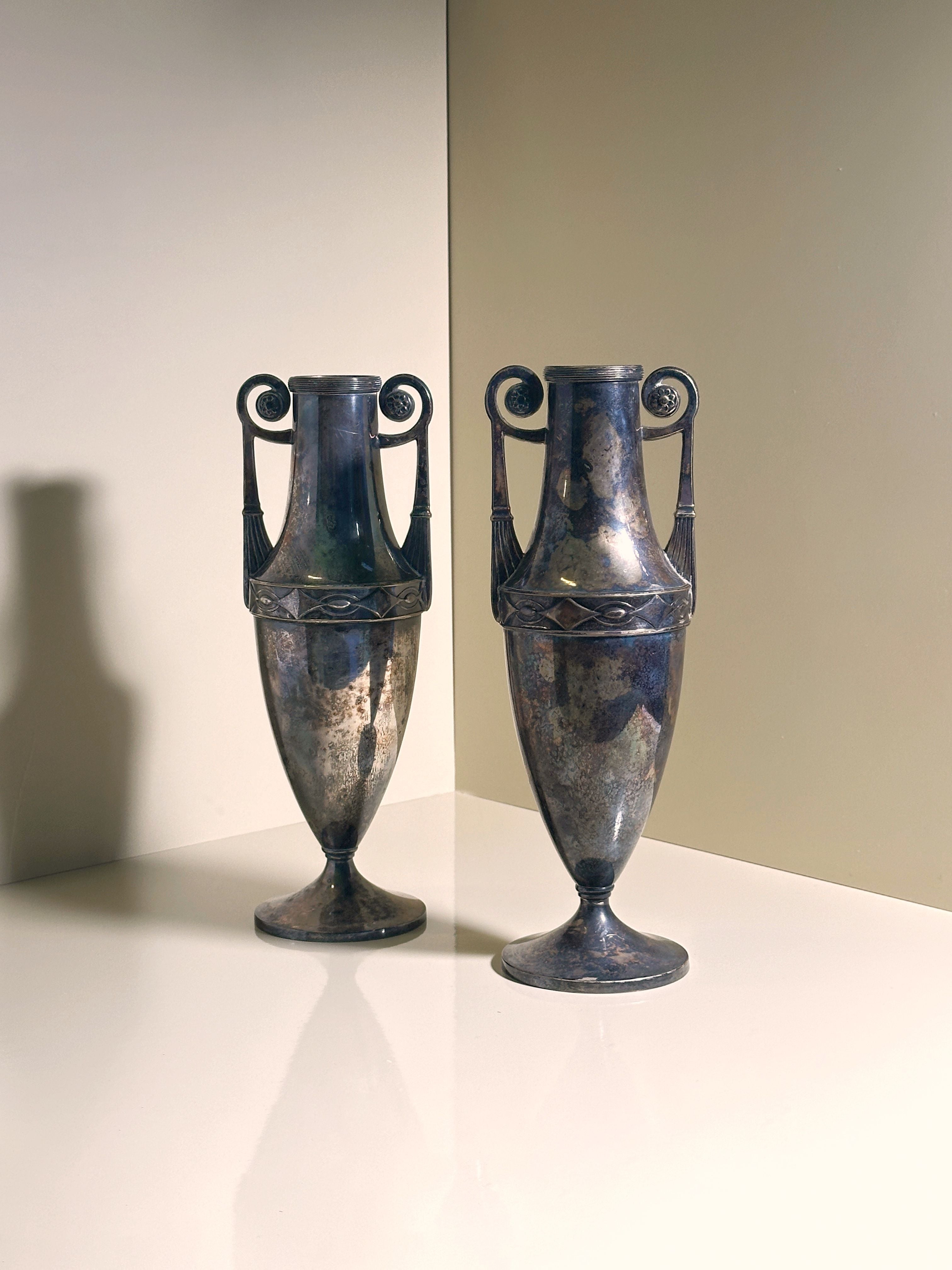 A pair of elegant, tall silver-plated Art Nouveau Amphora from 1890 by Dodo Vintage, with handles and intricate patterns, stand together on a smooth light surface against a neutral background, casting soft shadows.