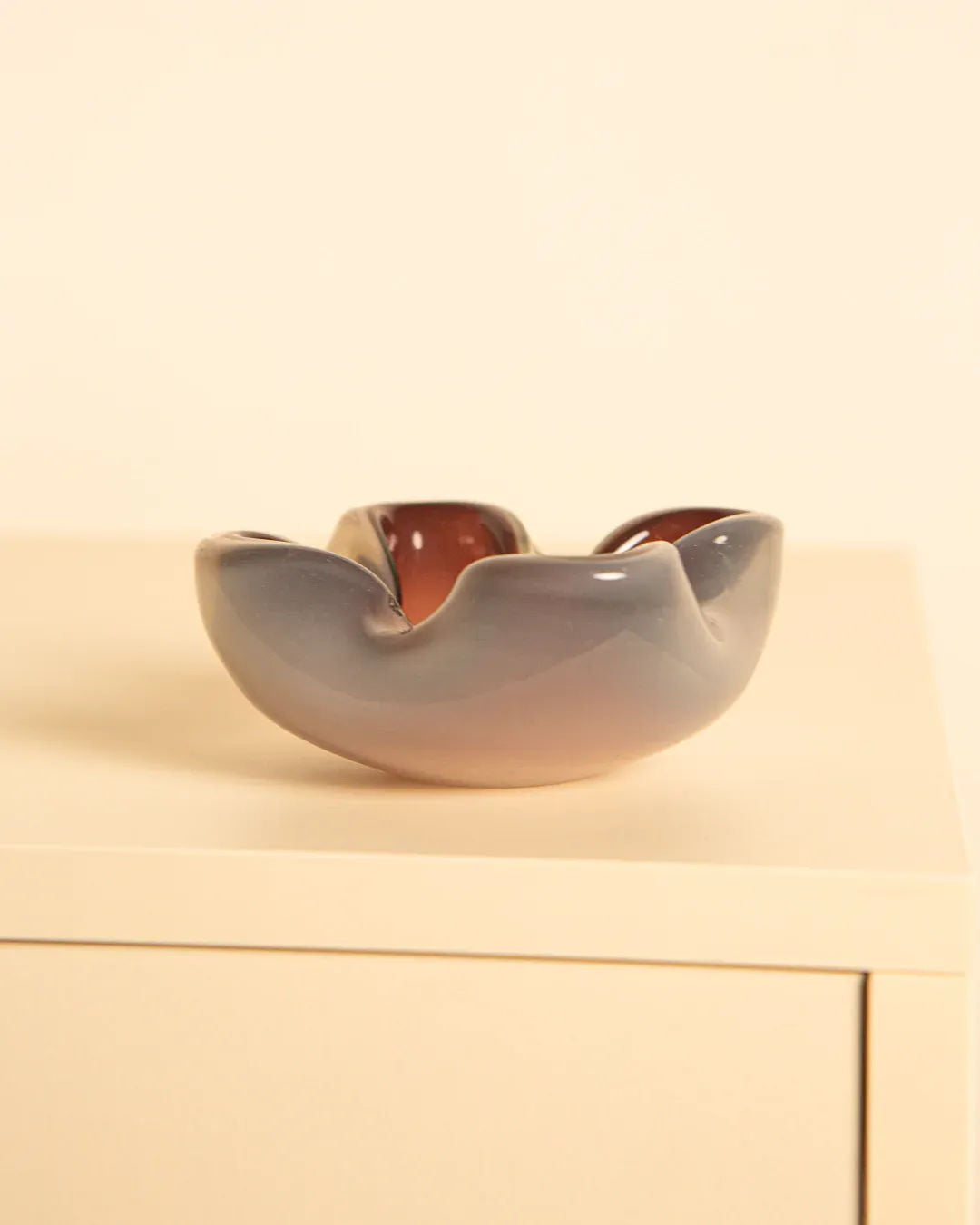 On a light-colored wooden surface rests Treaptyque's Murano Opaque Glass “Fleur” 60s bowl, featuring a smooth, wavy abstract shape reminiscent of vintage Italian design. With its gradient from muted pink to soft gray, the plain beige background accentuates its elegant allure.