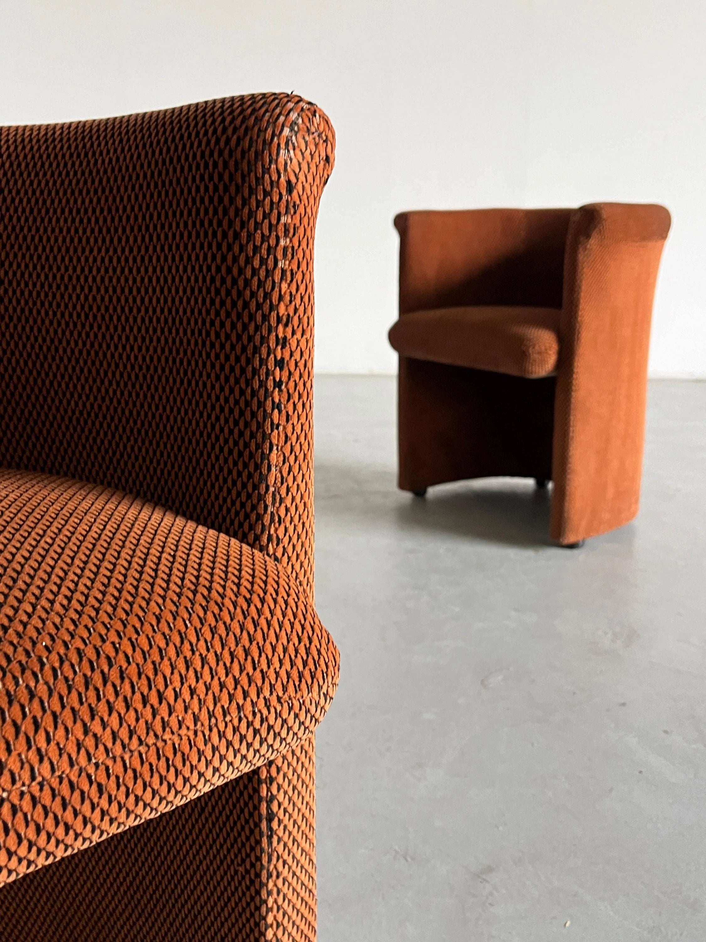 Close-up of Cherry Cargo's Pair of Postmodern Armchairs in vintage condition, showcasing orange geometric patterned fabric. Another identical chair rests on a concrete floor against a white wall, highlighting the classic design.